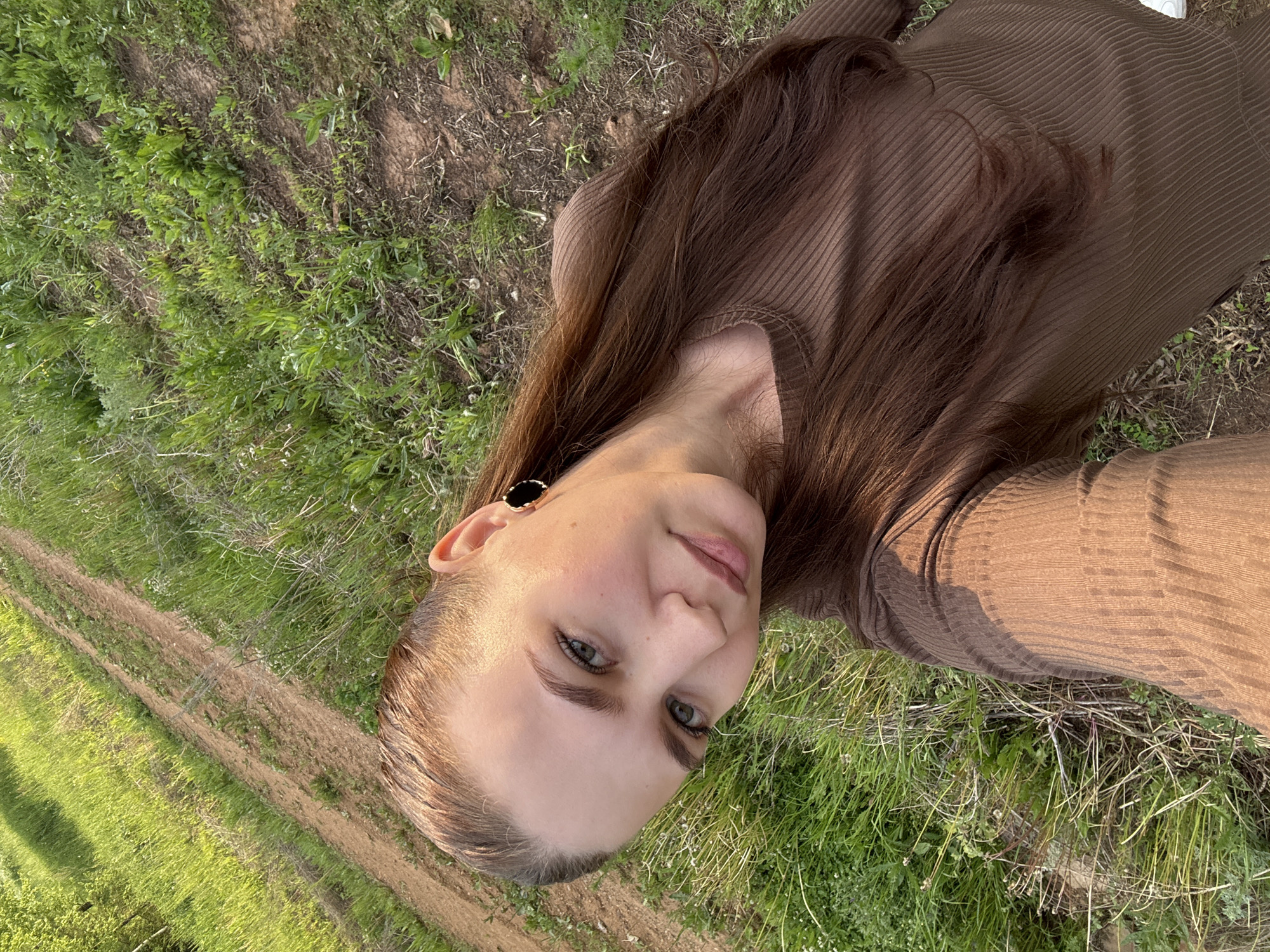 A young woman is captured in a close-up selfie or portrait, looking directly at the camera with a calm expression and a slight smile. Her head is tilted, giving the image a Dutch angle. She has long, straight, dark brown hair that cascades over her shoulder and is also visible spread around her on the ground. Her eyes are a striking green, and she has well-defined eyebrows and eyelashes. On her visible ear, she wears a black circular earring with a gold rim.
She is dressed in a brown, long-sleeved top or dress with a distinct ribbed texture. The setting is outdoors, likely a rural or semi-rural area in Nizhnyaya Gar', Russia. The background consists predominantly of vibrant green grass and patches of dry earth. A dirt path or track is visible curving through the grassy landscape, suggesting an open field or trail. The lighting appears to be natural daylight, indicating either morning or late afternoon, creating a soft glow on her skin and hair. There is no visible text in the image.