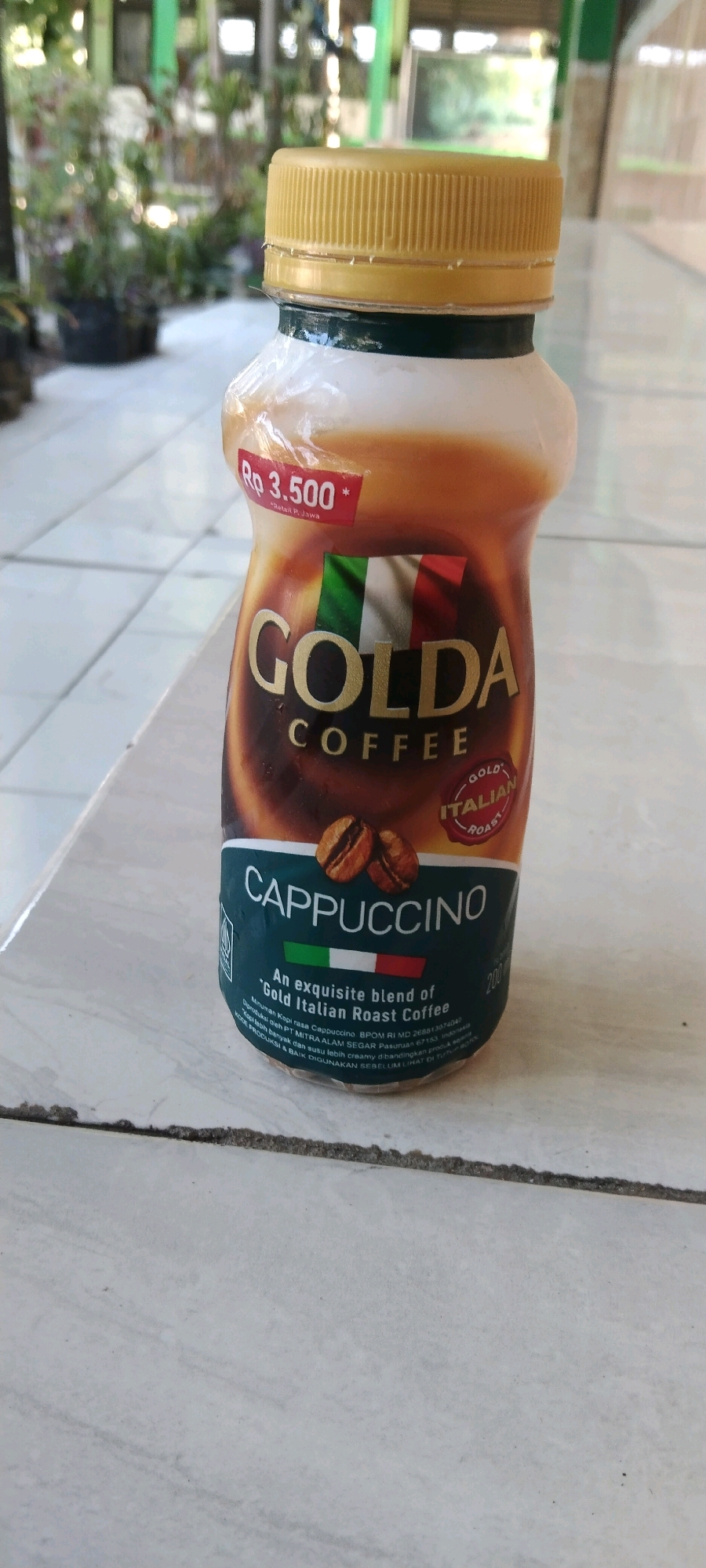 A bottle of Golda Cappuccino coffee drink, with a yellow cap, is placed on a tiled surface. The bottle has a label with "GOLD CAFE", an Italian flag, and the text "CAPPUCCINO" and "An exquisite blend of 'Gold Italian Roast Coffee'". A red sticker with "Rp 3.500" is also visible on the bottle. The background is blurred, showing greenery and a building structure, possibly outdoors in a semi-urban or residential area. The scene does not provide enough information to determine the specific location within Wadang, Indonesia.