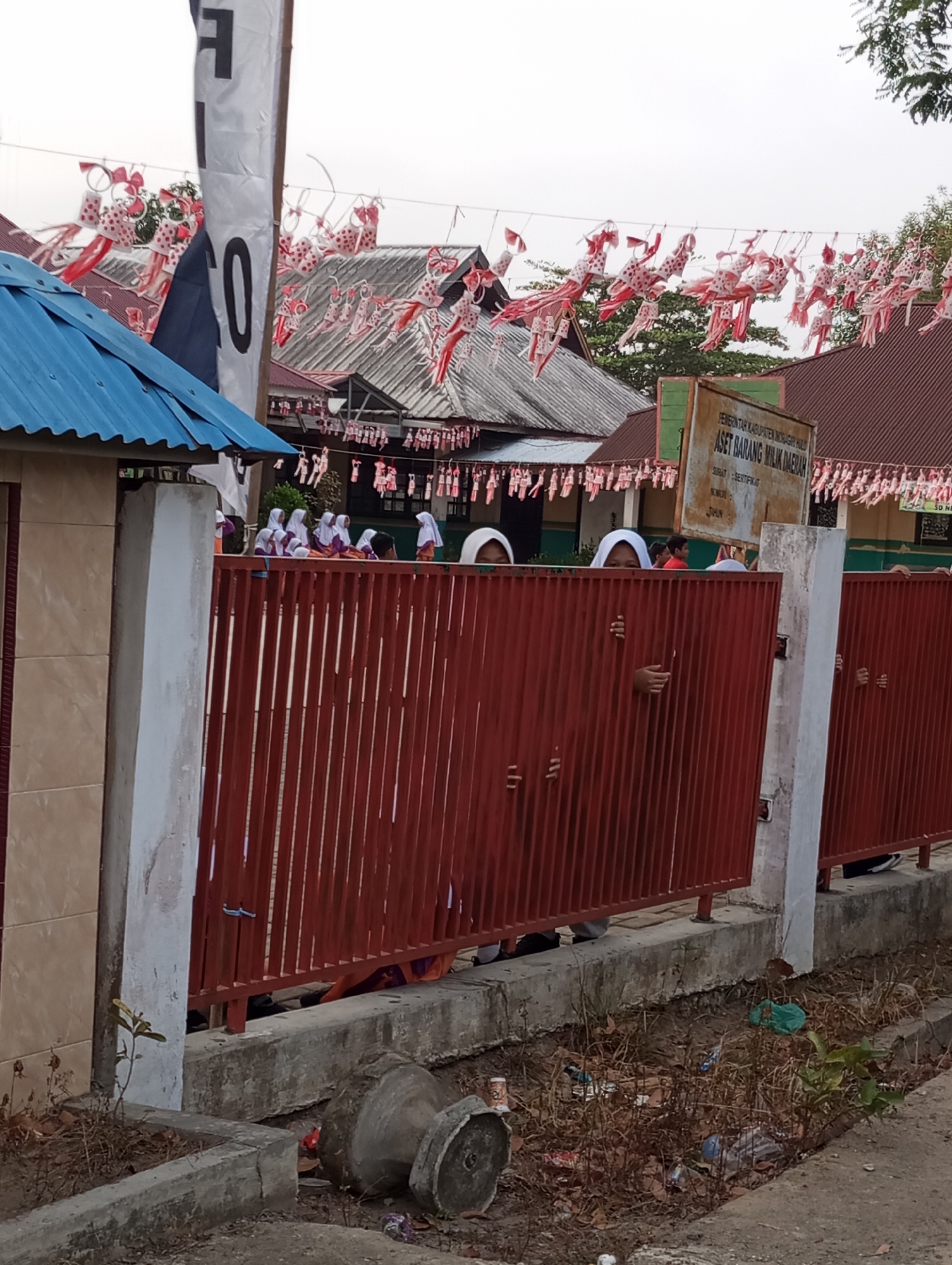 The image captures an outdoor scene in Pematang Reba, Indonesia, likely a school setting based on the presence of students. In the foreground, a red metal fence stretches across the frame, with several children's hands visible peeking through the bars. Behind the fence, a group of young people, predominantly girls wearing white hijabs, are gathered.
Above the fence, a tall white banner with black lettering is partially visible, with the number "10" prominently displayed. Numerous small, decorative flags or banners, predominantly red and white, are strung overhead, suggesting a festive or patriotic occasion. The background reveals buildings with grey tiled roofs, characteristic of Indonesian architecture. A weathered signpost is also visible, with text in Indonesian.
The environment appears somewhat neglected, with dry vegetation and scattered debris on the ground in the lower portion of the image, including broken concrete planters and various plastic items. The overall lighting suggests it is daytime, possibly late afternoon or early morning, with an overcast sky. The composition of the image focuses on the interaction between the children behind the fence and the surrounding environment.