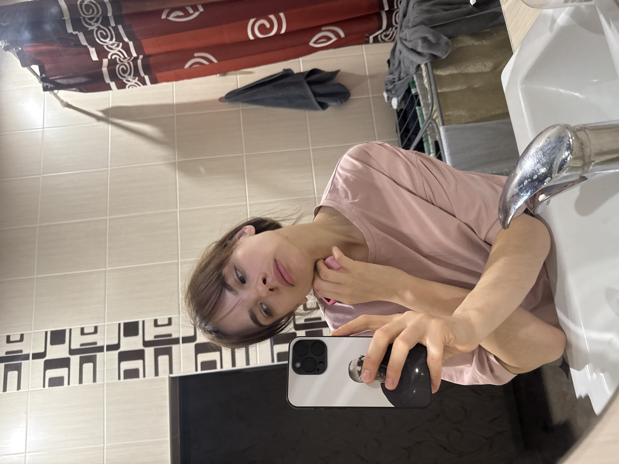 A young person, likely female, with light brown hair and light-colored eyes, is featured in a rotated selfie taken in what appears to be a bathroom in Rostov-on-Don, Russia. The image is oriented 90 degrees clockwise, so the subject's head is on the left and their body extends to the right.
The person is wearing a plain light pink t-shirt and looks directly at the camera with a neutral expression. They hold a white smartphone, which appears to be an iPhone with multiple cameras, in their left hand. The phone's back features a pop-socket or grip with a black and white graphic. Their right hand is raised to their chin, holding a small pink object, possibly a cosmetic item or beauty tool.
The background elements clearly indicate a bathroom setting. To the upper left of the rotated image, a red shower curtain with distinctive white circular patterns is visible. The wall behind the person is tiled with light beige square tiles, featuring a horizontal decorative band of darker brown and beige geometric patterns. To the right, a white sink with a reflective chrome faucet is prominent. Further in the background, visible through a partial reflection in a mirror, is a laundry hamper or a drying rack with clothes on it, and a dark grey towel is folded on the tiled floor. The lighting is artificial, typical of an indoor environment. There is no legible text visible in the image.