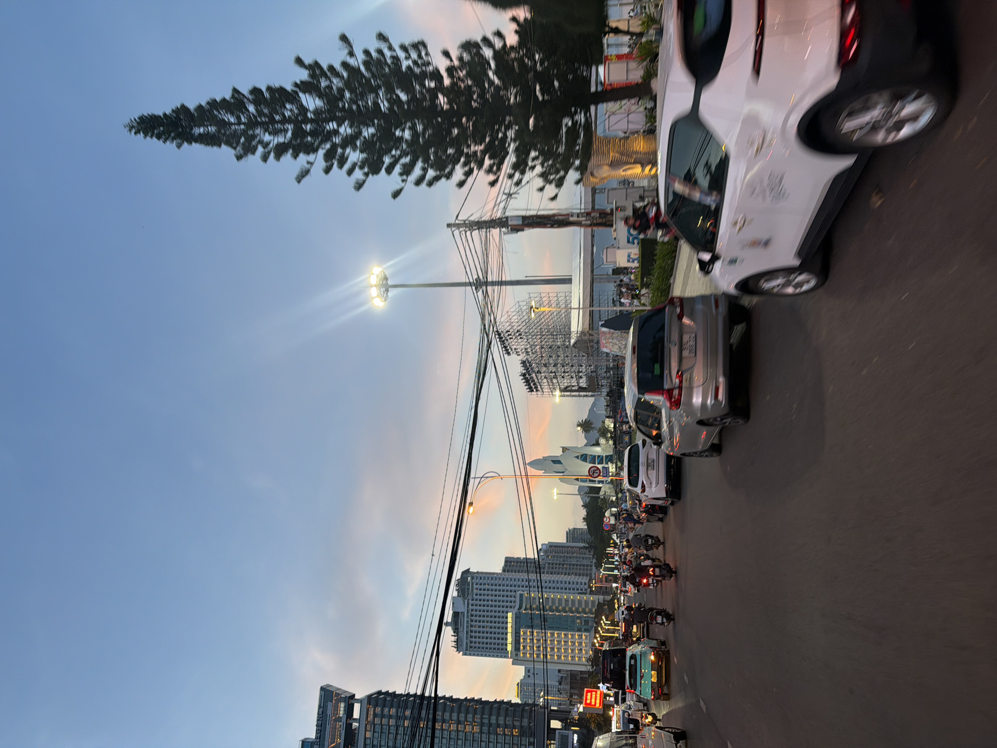 The image captures a busy street scene in Nha Trang Ward, Vietnam, during what appears to be twilight or early evening. The sky exhibits a soft gradient of blue and pastel pink, indicative of sunset or sunrise. A dense traffic flow dominates the foreground, featuring a mix of cars and numerous motorcycles, many carrying multiple passengers. The vehicles are on a paved road, with lanes clearly marked by painted lines.
Several tall buildings, characteristic of an urban environment, line the left side of the street. One prominent building on the far left displays the word "REGENCY" on its facade. Other skyscrapers, some illuminated by interior lights, are visible further down the street. On the right side of the road, a tall, slender evergreen tree stands out against the sky, its silhouette dark and distinct. A tall light pole with multiple illuminated lamps is also visible, casting beams of light.
Further down the street, a distinctive building with a pointed, white, spire-like structure can be seen. Structures that resemble scaffolding or unfinished buildings are also present in the background, suggesting ongoing construction or event setup. Power lines are strung across the scene, contributing to the urban atmosphere. The overall impression is one of a vibrant, active city street with a significant amount of vehicular and pedestrian activity.