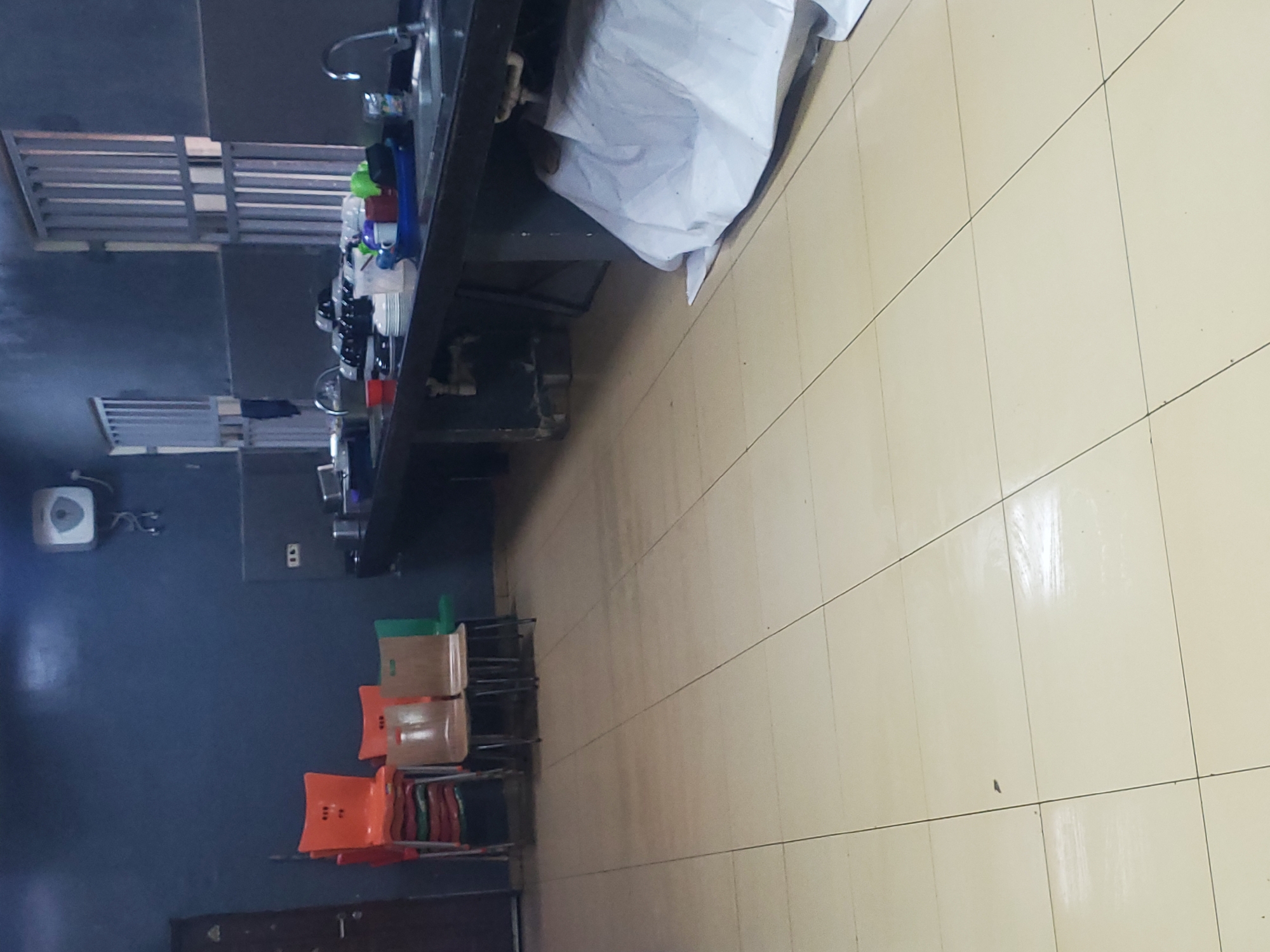 The image depicts a dimly lit room, possibly a communal kitchen or dining area, located in Gwagwada, Nigeria. The floor is tiled with light-colored squares, showing some wear and tear. To the left, a stack of orange chairs is visible, with a few wooden chairs placed beside them. A dark-colored door is partially visible on the far left.
The main focus of the scene is a long, dark countertop, likely a kitchen counter, running across the middle and right side of the image. It is cluttered with various kitchen items. On the counter, there are stacks of plates, cups, and other dishes, some appearing to be clean and neatly arranged, while others are in a blue basin, suggesting they are in use or waiting to be washed. A faucet and sink are visible on the right side of the counter. Behind the counter, a dark wall with barred windows suggests the building might be secure.
A large white sheet or cloth is draped over a section of the counter or furniture underneath it on the right side, partially obscuring what is beneath. In the upper left corner of the image, a white unit, possibly a water heater or air conditioning unit, is mounted on the dark blue wall. The overall impression is one of a functional, albeit somewhat unkempt, common area.