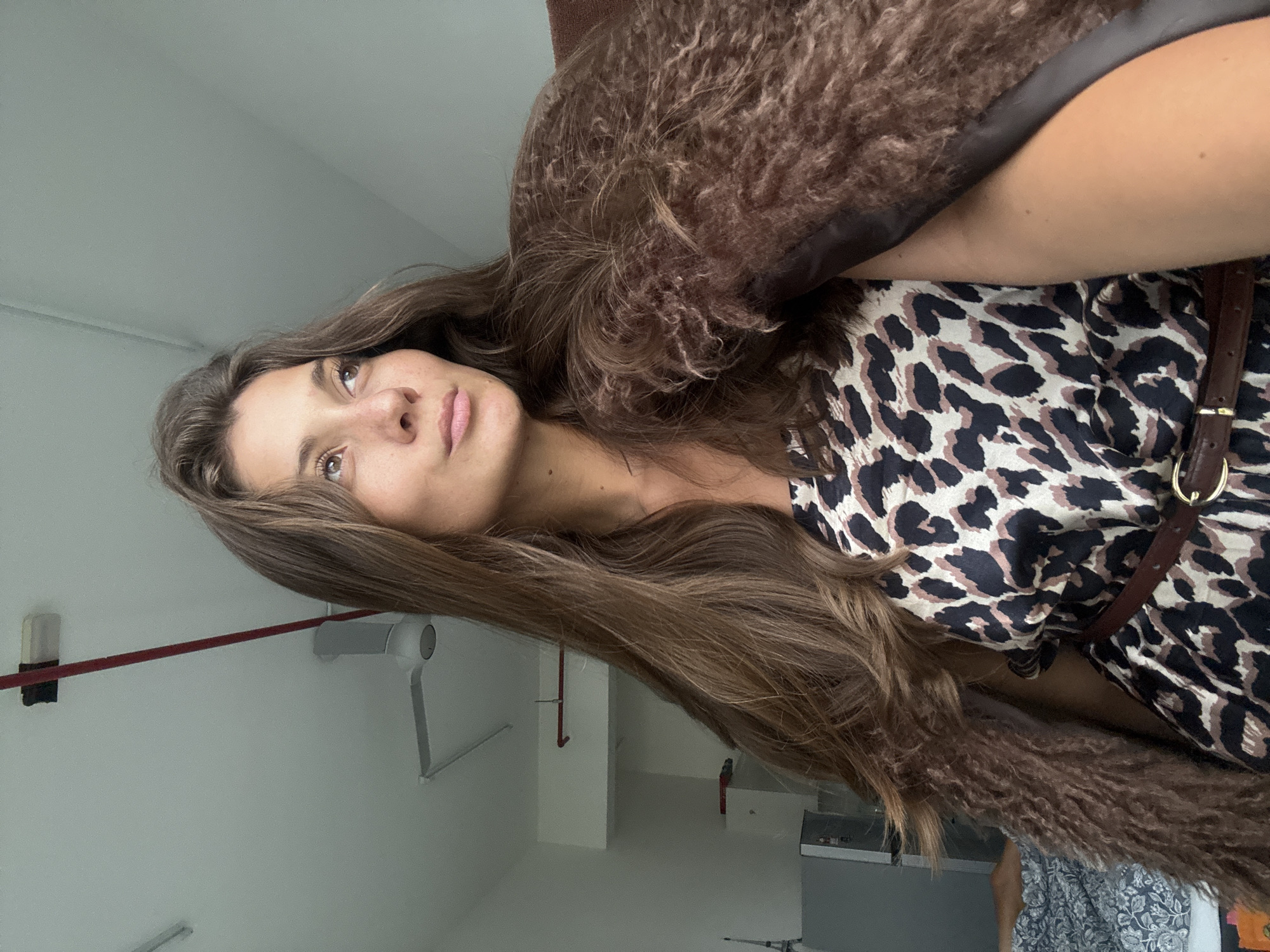 The image shows a woman with long brown hair and a leopard-print dress. She is wearing a brown belt with a gold buckle. The background is a white wall with some pipes and a device hanging on the wall. There is also some furniture in the background. This image does not provide sufficient information to determine the exact location in Kuala Lumpur, Malaysia, or any specific actions or interactions occurring.