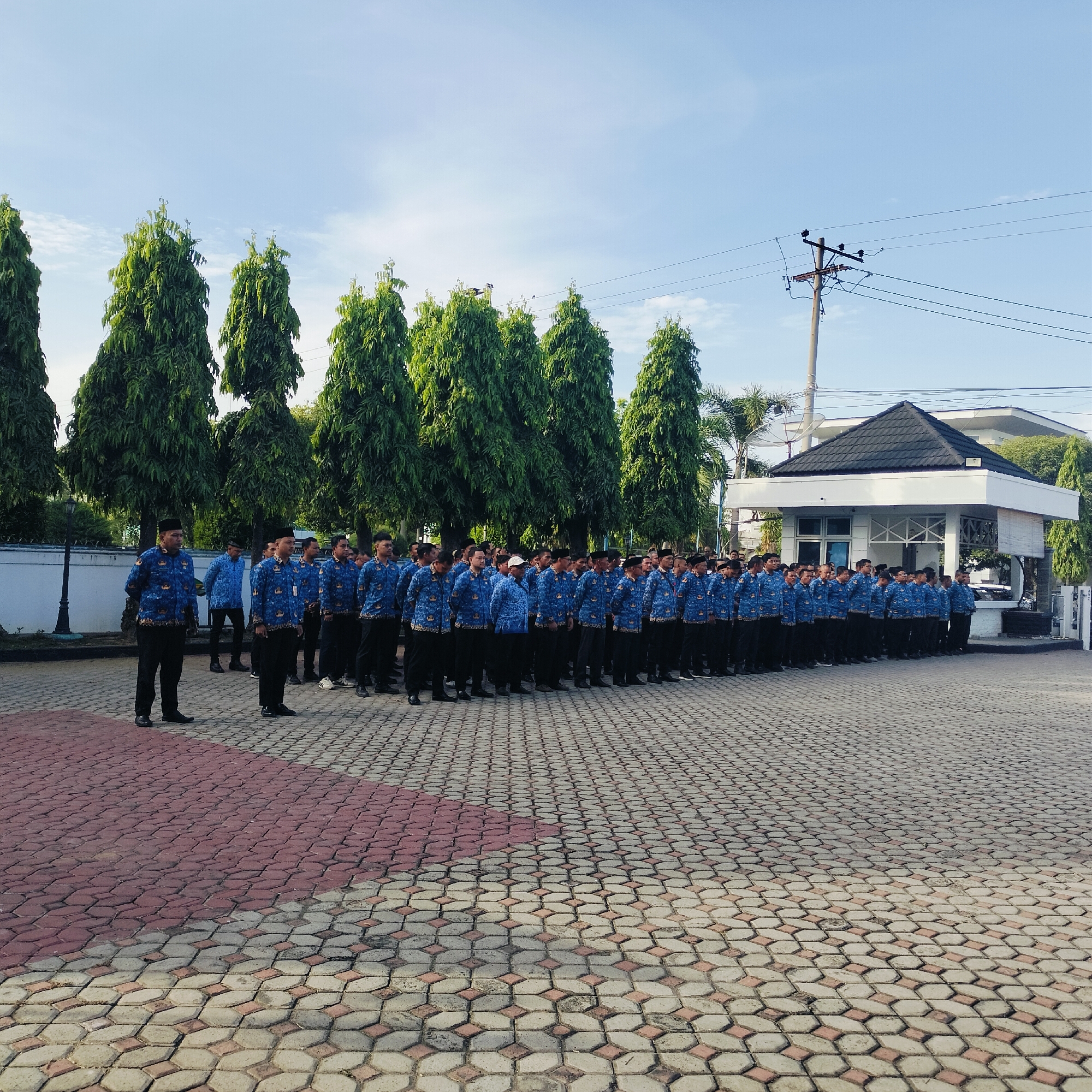 A large group of men in blue patterned shirts and black pants are standing in formation outdoors in Lhokseumawe, Indonesia. They are arranged in neat rows on a paved surface with a red and gray honeycomb pattern. Tall, evergreen trees line the perimeter behind them, with a white building featuring a black-tiled roof in the background. A utility pole with wires is visible near the building. The sky is blue with sparse clouds. The men appear to be participating in some sort of gathering or event.