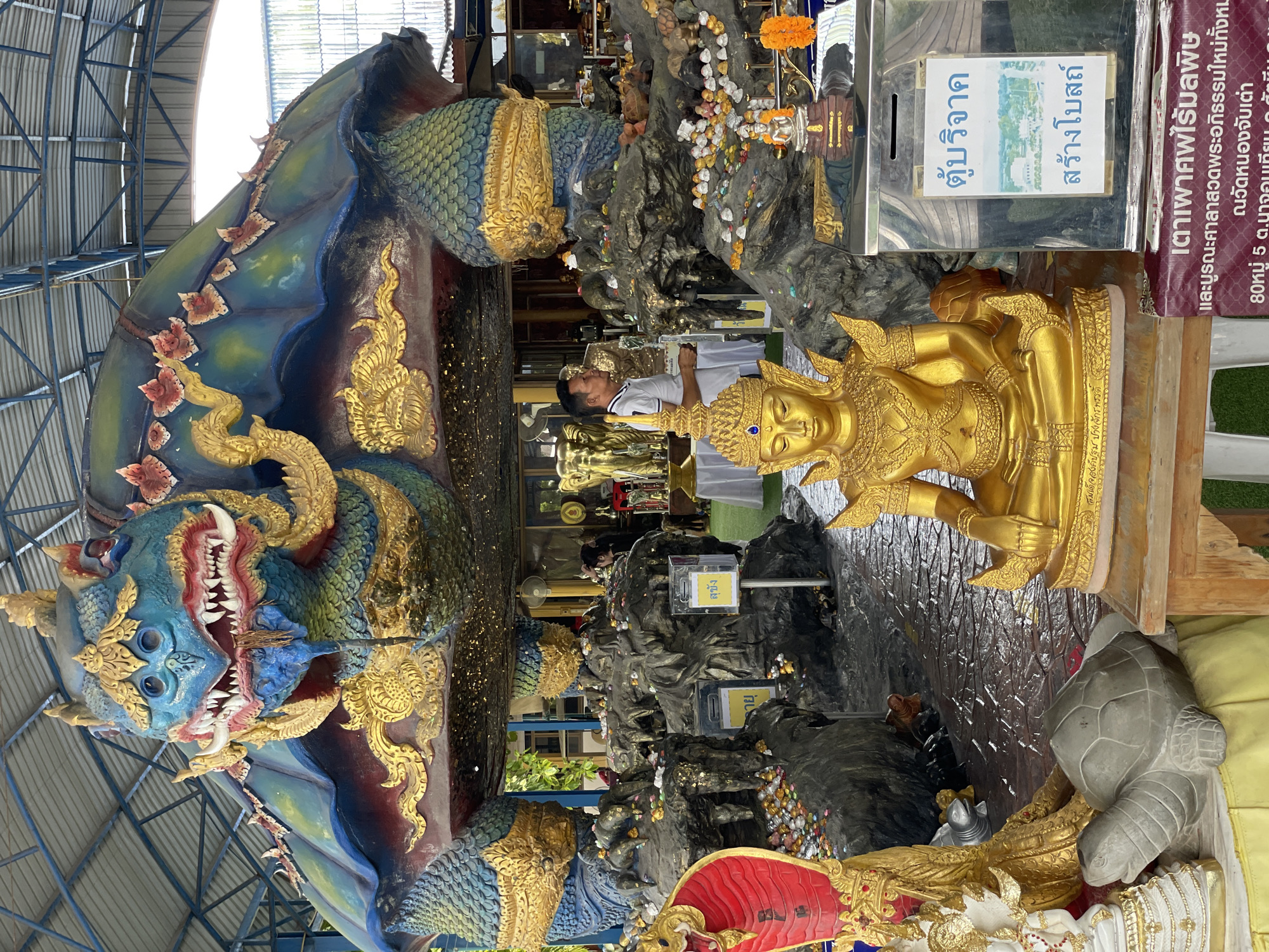 The image captures an outdoor scene within a covered structure, likely a temple or shrine, in Bang Sare, Thailand. Dominating the upper half of the frame is a colossal, vibrantly colored statue of a mythical sea turtle. Its head is detailed with a blue face, red and white fangs, and golden accents. The turtle's shell is a mosaic of blues and greens, adorned with intricate golden patterns and floral motifs.
Beneath the massive turtle, the environment is cluttered with numerous statues and offerings. In the foreground, a golden statue of a seated Buddha, adorned with a golden crown, is placed on a wooden platform. To its right, a metal donation box with a white sign featuring Thai text and an image of a building is visible. A sign in Thai language with red lettering is placed below the donation box. To the left of the Buddha statue, a stone turtle sculpture lies on a yellow cloth. Further back and to the left, a tall statue of a red cobra rears its head.
The ground is paved with patterned stones, appearing wet in places, suggesting recent rain or cleaning. The middle ground is filled with rocky formations, upon which are scattered numerous small offerings, including coins and colorful pebbles. A man in a white polo shirt stands to the right of the rocky formations, looking towards the left. In the background, glimpses of other golden statues, including elephants, and more offerings are visible, along with parts of buildings and foliage. The entire scene is covered by a corrugated metal roof supported by metal beams, suggesting an open-air pavilion. The lighting is diffused, indicating an overcast or shaded environment.