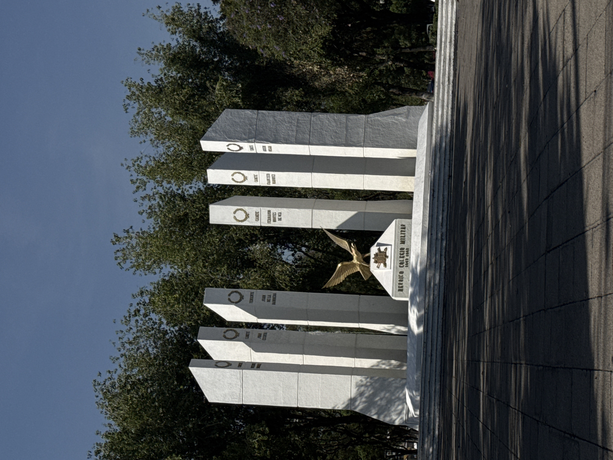 The image depicts a monument honoring military heroes in Puebla, Mexico. The monument consists of several white, rectangular pillars arranged in ascending order, each bearing the name of a military hero. A golden eagle is affixed to a central pillar. In the background, the monument is framed by lush green trees. The monument is set on a paved surface, with shadows cast by trees. The sky is visible in the upper left corner.