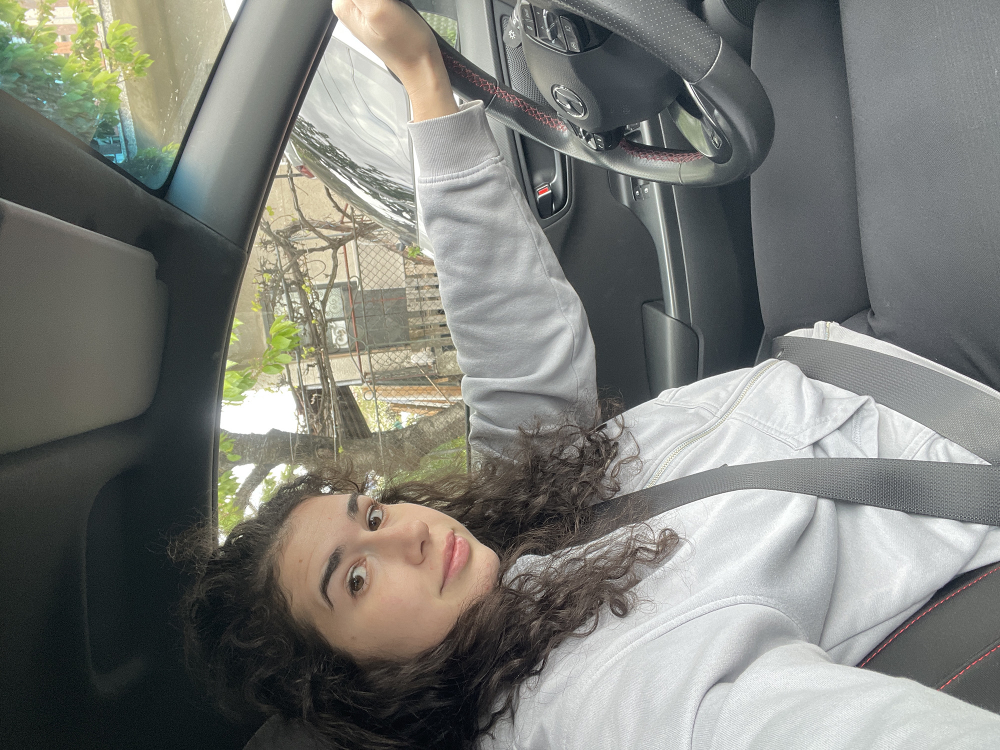 A young woman with curly dark hair is inside a vehicle. She is wearing a light gray hooded sweatshirt and a seatbelt. Her left hand is on the steering wheel. The interior of the car is visible, including the steering wheel with red stitching, dashboard, and seats. Outside the car, there are trees and a building. The location is presumably Sofia, Bulgaria.
