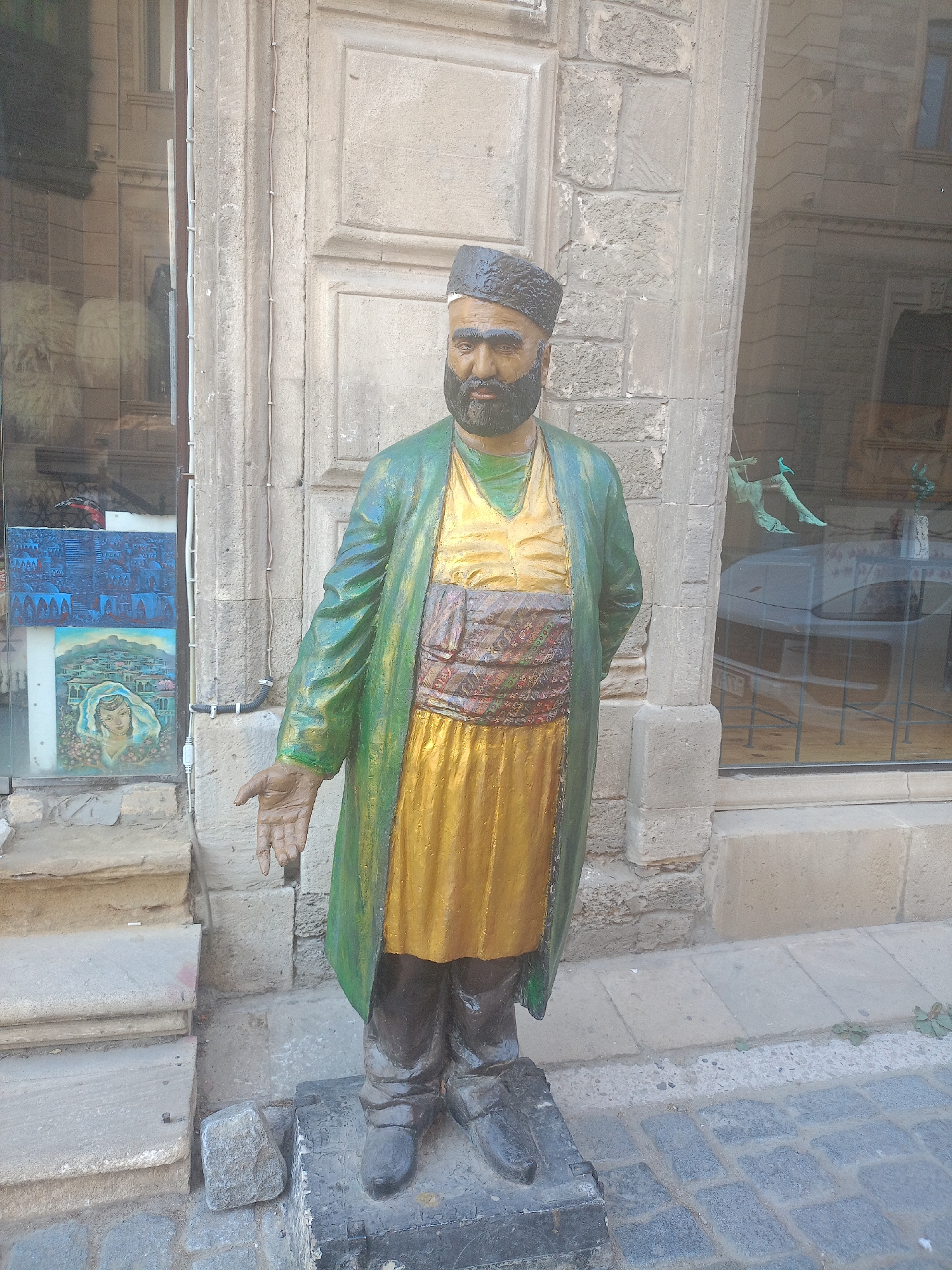 A full-body, slightly low-angle, eye-level shot shows a weathered, life-sized statue of a man in traditional clothing, standing on a stone pedestal on a cobblestone street. The man wears a black patterned cap, a green outer robe over a gold tunic, and a red, patterned sash around his waist. He has a beard and is shown with his left hand outstretched, palm up, as if to receive something. The statue stands in front of a building with rough stone walls and large windows. In the background, through the window, a table and chairs can be seen, as well as what appears to be a car parked behind them. To the left of the statue, a set of stone stairs leads up to a shop entrance, where two paintings are displayed in the window. The lighting suggests it is daytime, with shadows indicating a sunny environment.