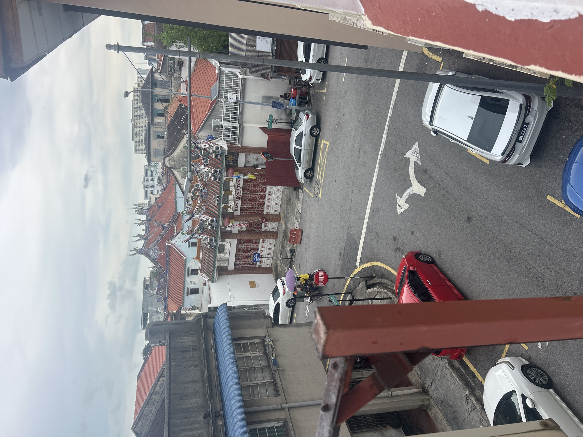 The image captures a street scene in George Town, Malaysia, likely on a cloudy day. In the foreground, a red metal railing partially obstructs the view. To the left, a white car is parked on a street with a yellow line. Further down the street, a red car is also parked. In the center of the frame, a white car is parked, and beyond it, a silver car is visible. A person is standing near a blue tub beside a building with the sign "KEW LEONG TONG 1980". The street is paved with asphalt and marked with white lines. Directly in front of the "KEW LEONG TONG" building, a red gate leads into a courtyard with ornate roof decorations. Various residential and commercial buildings with terracotta tile roofs are visible in the background, with a taller modern building in the distance. A street light is present on the right side of the image.