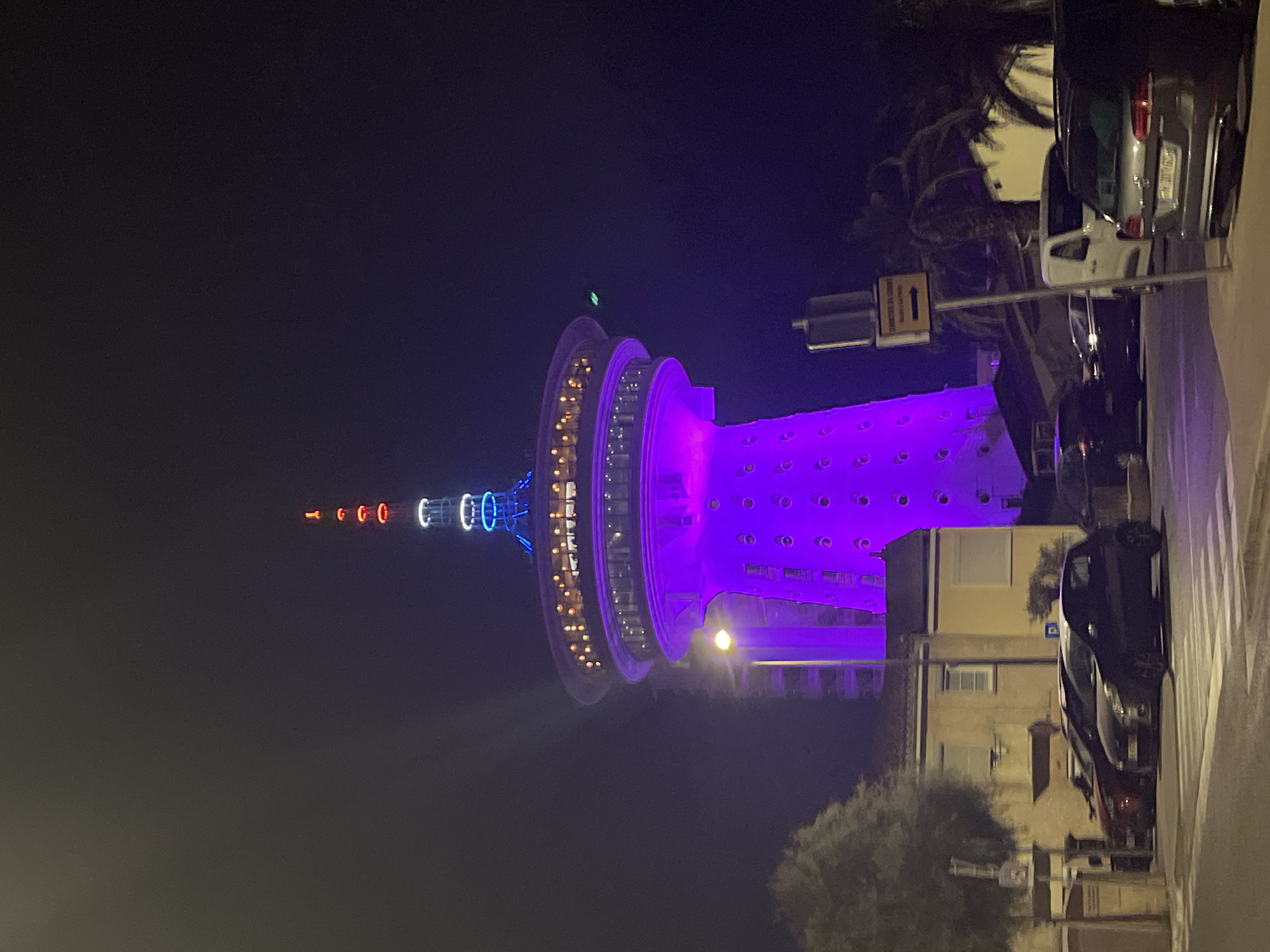 The image shows the Phare de la Méditerranée, a lighthouse and observation tower, at night in Palavas-les-Flots, France. The tower is illuminated with purple lights at its base and blue lights on the upper sections, with red and white lights at the very top. There is a circular structure with illuminated windows partway up the tower, suggesting it might house a restaurant or other facilities. In the foreground, several cars are parked on a street, with a sidewalk and a tree visible. The overall environment appears to be an urban setting at night, with the tower being the dominant feature.