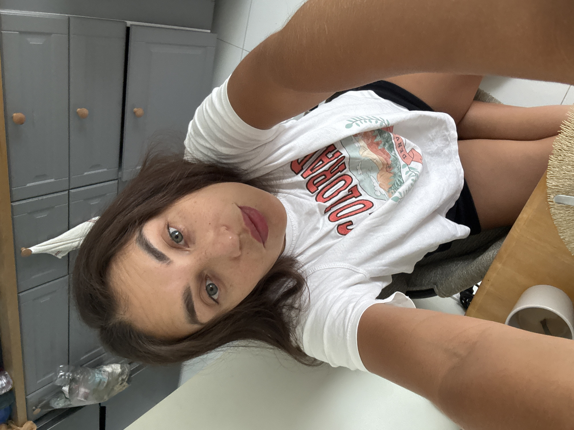 A selfie photograph shows a woman with shoulder-length brown hair, fair skin, and green eyes. She is wearing a white t-shirt with a graphic design and red lettering. She appears to be indoors, possibly in a bathroom or closet area, as grey cabinet doors with light brown knobs are visible in the background. Other items such as a toothbrush and a table with a grey towel on it can also be seen in the image. There is nothing in the photograph that suggests this image was taken in Pa Khlok, Thailand.