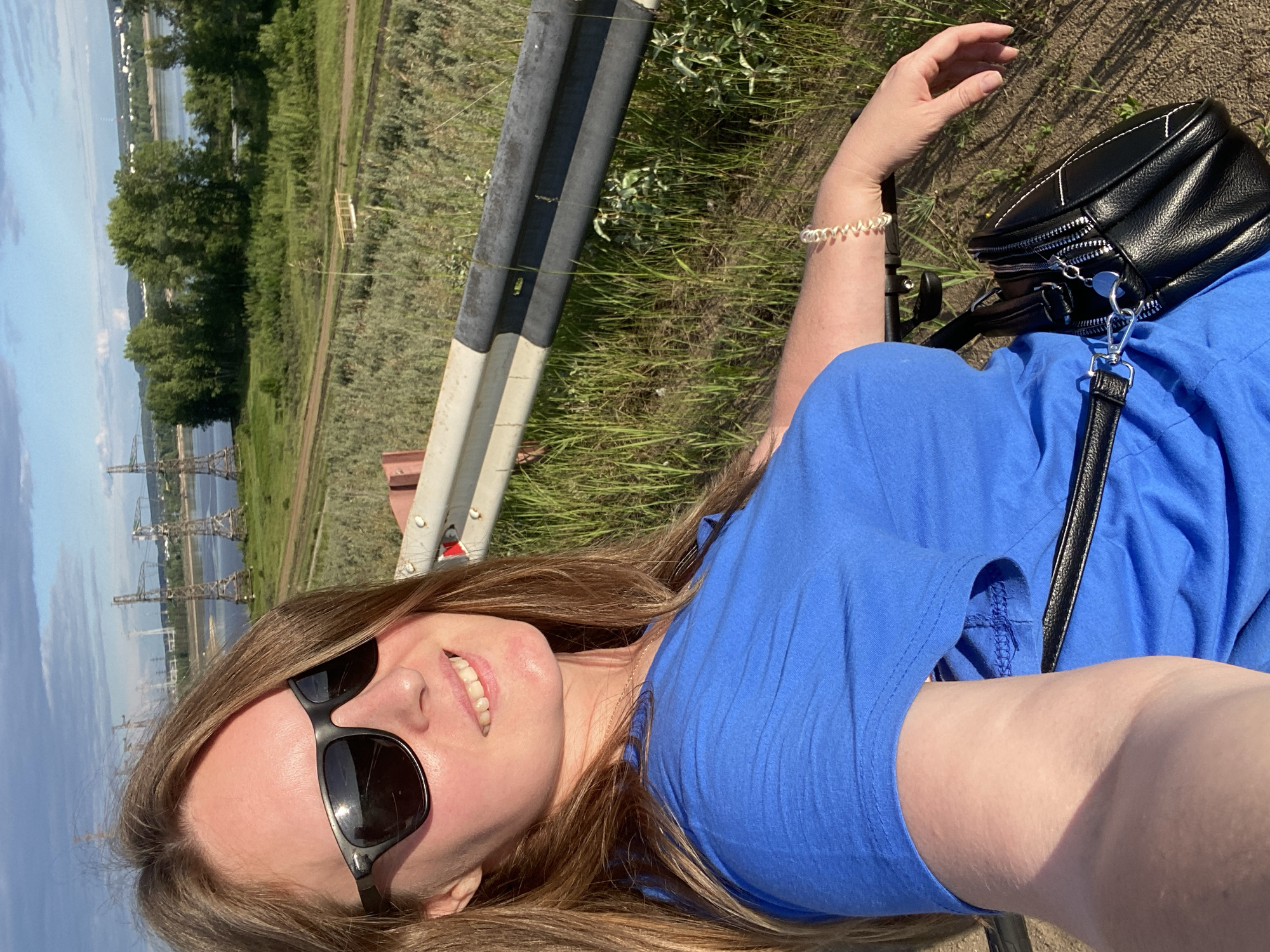 A female individual, appearing to be an adult, is depicted in a selfie-style shot outdoors, positioned in front of a natural landscape featuring a river and distant industrial infrastructure. The setting is in Chaikovsky, Russia.
The woman has long, light brown hair and is wearing black sunglasses, a bright blue short-sleeved top, a thin gold-colored necklace, and a light-colored spiral-shaped bracelet on her right wrist. She is smiling, revealing her teeth. A small black handbag with multiple silver zippers and a shoulder strap is visible across her torso. Her skin appears fair, with some areas exhibiting a slight redness, indicative of sun exposure.
Immediately behind the woman is a black and white metal guardrail. The foreground and midground feature dry earth, green grass, and various types of green vegetation, including bushes and trees. A narrow dirt path is visible in the middle ground, running parallel to a wide body of water identified as a river. A small, light-colored wooden bridge or structure spans a section of the path or a small channel.
In the background, across the river, several large, lattice-structured power transmission towers are prominently visible. Beyond the river and towers, a distant cityscape or settlement with buildings and a tall antenna or tower can be discerned on the horizon under a sky with scattered white clouds. The lighting suggests it is daytime.