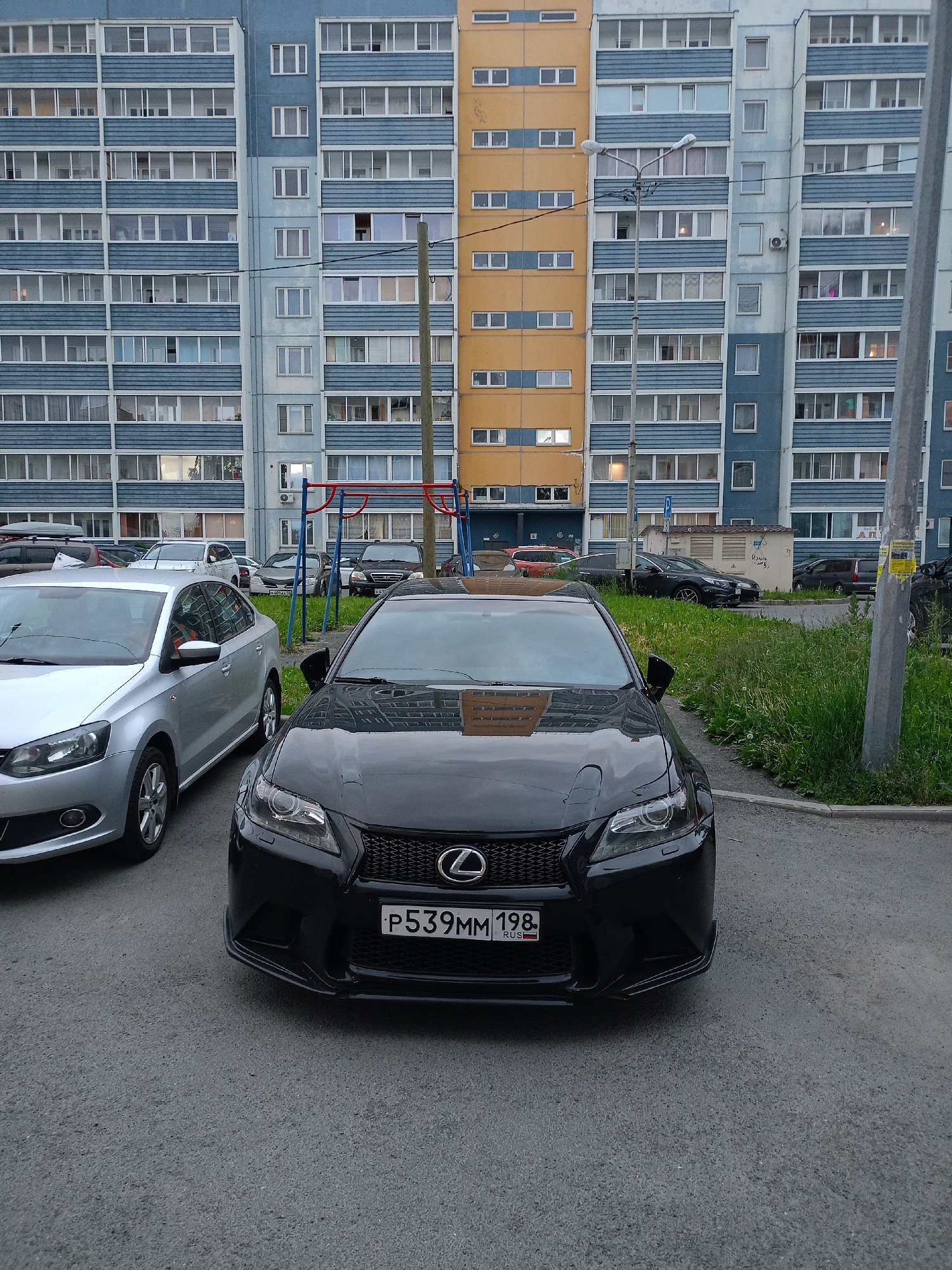 A black Lexus sedan is parked on asphalt in front of a multi-story residential building in Petrozavodsk, Russia. The building has a blue and white facade with numerous windows and balconies. The car's license plate reads "P539MM 198 RUS". To the left of the Lexus, a silver Volkswagen Polo sedan is parked. The ground is a mix of asphalt and green grass. A swing set is visible in the background.