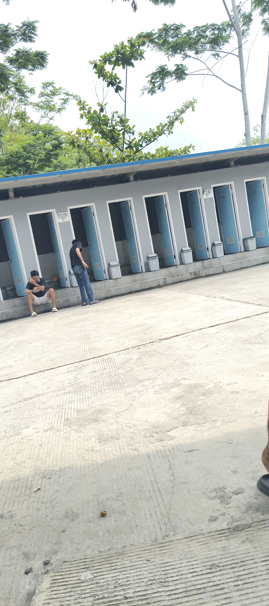 The image depicts an outdoor area with a row of public restrooms. Two individuals are present. One person, a male wearing a dark t-shirt and white shorts, is sitting on a concrete step facing away from the camera, looking down at a mobile phone. The second person, also male, is standing to the right of the first, wearing a dark t-shirt and blue jeans, with his back mostly to the camera. The restrooms are modular units with blue doors, some of which are open, revealing interior stalls. Trash cans are placed in front of the restrooms. The ground is paved with concrete, showing visible expansion joints and some wear. Trees and foliage are visible in the background, with a bright sky overhead, suggesting a daytime scene. The location is identified as Bukateja, Indonesia.