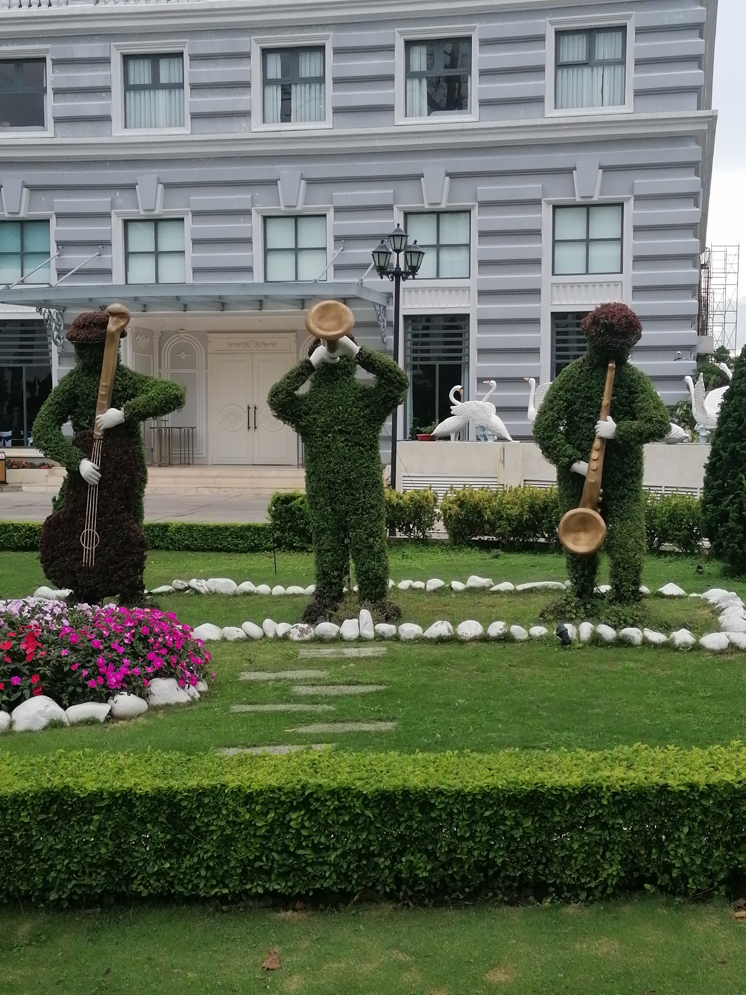 The image shows a garden with three topiaries shaped like musicians. The topiaries are green and are positioned on a grassy lawn. One topiary on the left holds a cello-like instrument. The middle topiary is playing a trumpet. The topiary on the right is holding a saxophone. In the background, a building with classical architecture and windows can be seen, along with some white swan sculptures. Pink and red flowers are planted in a bed to the left of the topiaries, bordered by white rocks. The scene is located in Dalat, Vietnam.