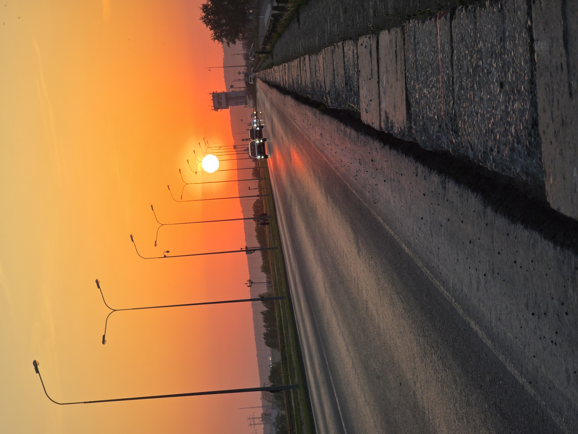 The image shows a road in the city of Chaikovsky, Russia, at sunset. The sky is a gradient of orange and yellow, with the sun low on the horizon. Streetlights line the side of the road. Several cars are traveling on the road, their headlights visible. A concrete barrier runs along the right side of the road. In the distance, a transmission tower and some trees are visible.