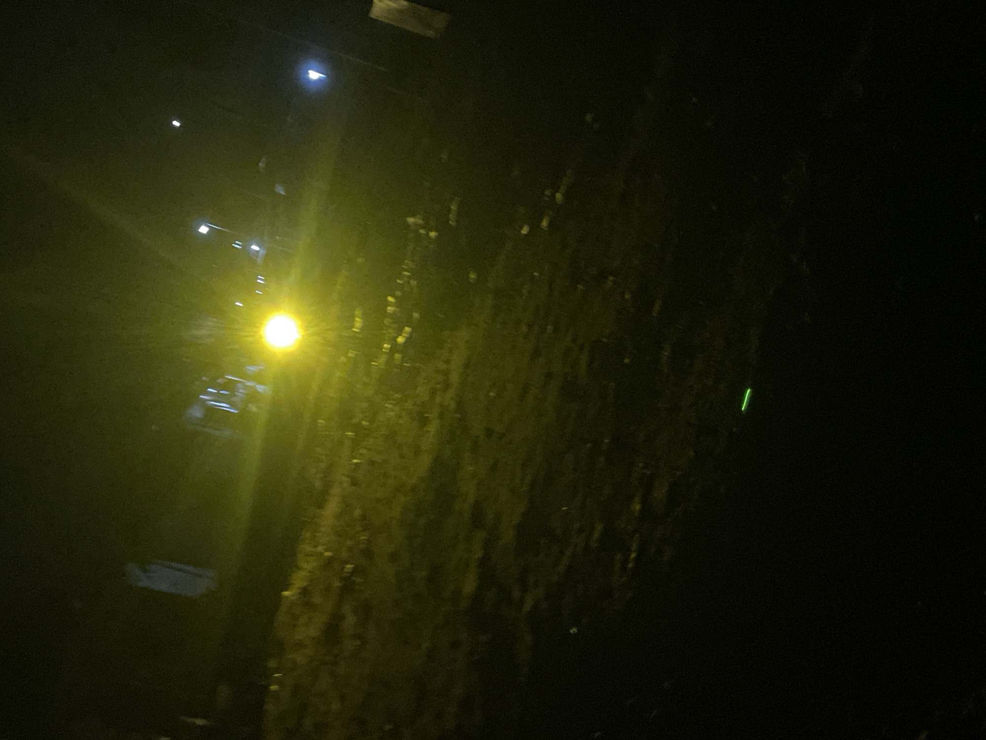 The image depicts a dark, outdoor scene at night, illuminated by artificial lights, likely in Lokoja, Nigeria. A bright, yellowish light source dominates the upper left portion of the image, casting an orange glow over the surroundings. In the mid-ground, there are several indistinct vertical structures that appear to be streetlights or poles, some with smaller, brighter lights at their tops. The ground in the foreground is dark and textured, possibly indicating soil or rough terrain. There is a small, bright green line visible in the lower center of the image, which could be a reflection or an artificial light source. The overall impression is of a dimly lit urban or semi-urban environment at night, with limited visibility and a dominant yellow cast from the primary light source.