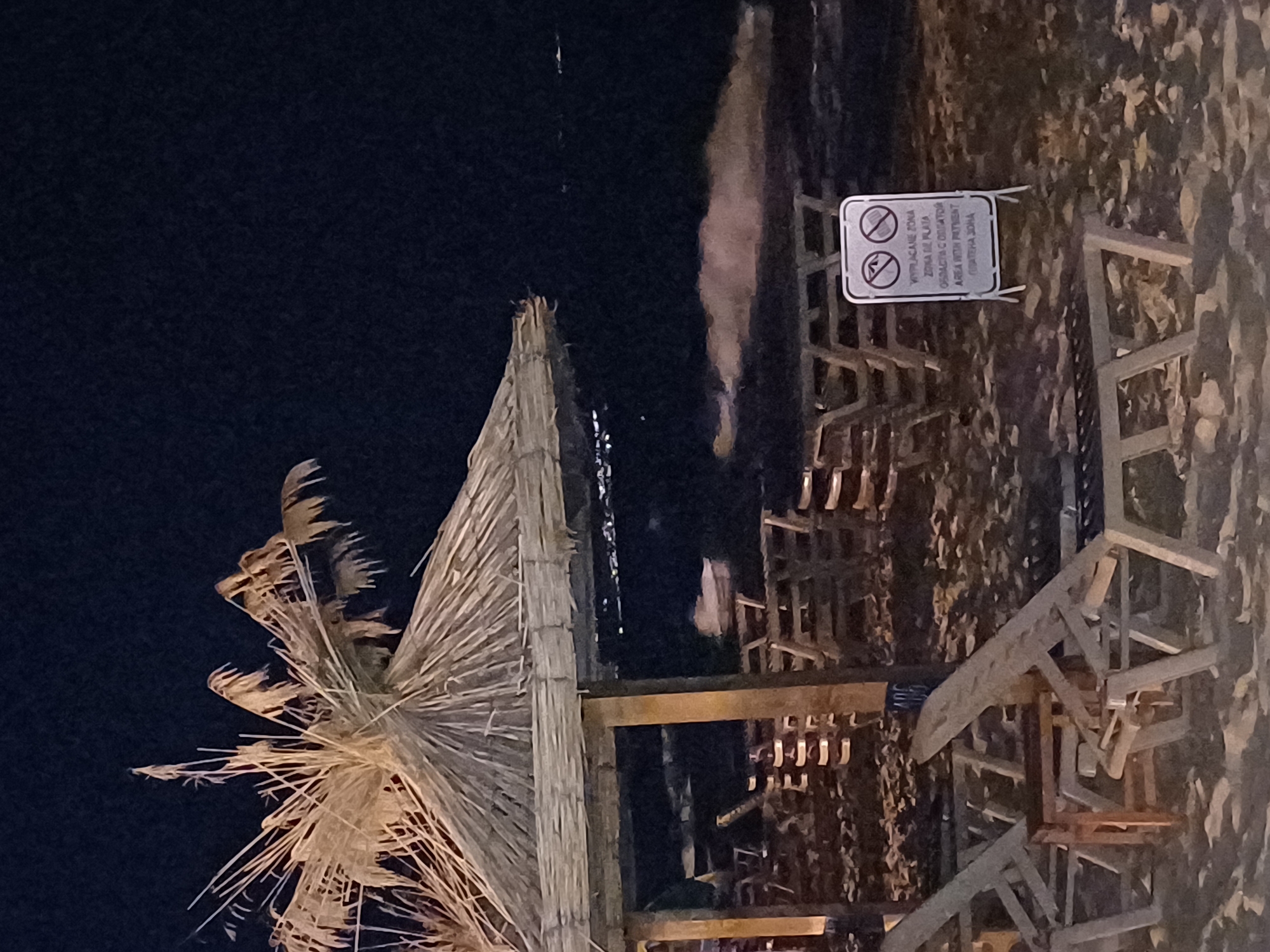 The image depicts a nighttime scene on a sandy beach in Varna, Bulgaria, near a thatched umbrella and several empty lounge chairs. In the background, the sea is visible with distant lights on the shore. A sign in the foreground reads "WYPRZEDAWANE ZONA ZONA NE PLATA OGRADASCTA C OBLIGATOR AREA WITH PAYMENT OBATENA ZONA."