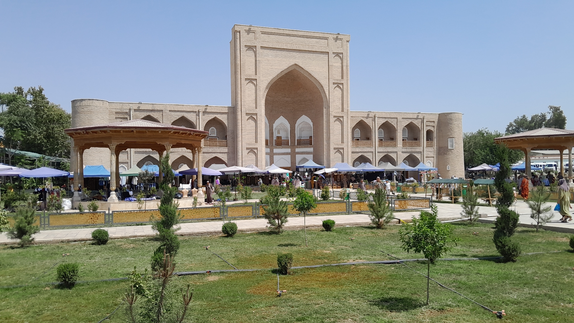 The image depicts a bustling outdoor market scene in front of a large, ancient-looking beige brick building with arched doorways and windows. The building appears to be a historical monument or religious site. In the foreground, a manicured lawn with scattered evergreen shrubs and trees is visible. A pathway runs through the lawn, bordered by a decorative fence.
The market itself is set up with numerous stalls covered by colorful tents, primarily blue and white, displaying various goods. People are seen browsing, shopping, and interacting within the market area. Some individuals are wearing traditional Uzbek clothing. To the left, a wooden gazebo with an ornate roof and columns offers shade.
The overall environment is sunny with a clear blue sky. Trees line the perimeter of the market and the historical building. The architecture of the building suggests a historical significance, possibly a madrasah or mosque, characteristic of the region of Denov, Uzbekistan. The presence of market stalls indicates a vibrant local economy and cultural activity.