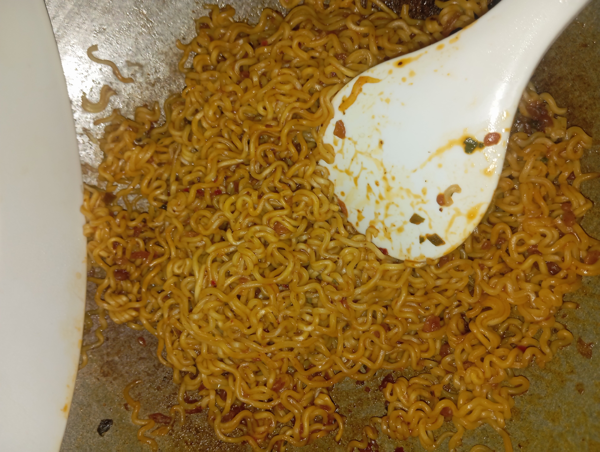 The image shows a close-up overhead view of instant noodles being cooked in a pan. A white spatula is partially obscuring the noodles on the right side of the frame. The noodles are curly and appear to be coated in a sauce with visible bits of chili and possibly other ingredients. The pan itself is dark and appears to have a seasoned, slightly oily surface. The overall scene suggests a domestic cooking environment, likely in a kitchen in Rejosari, Indonesia. There are no people visible in the image.