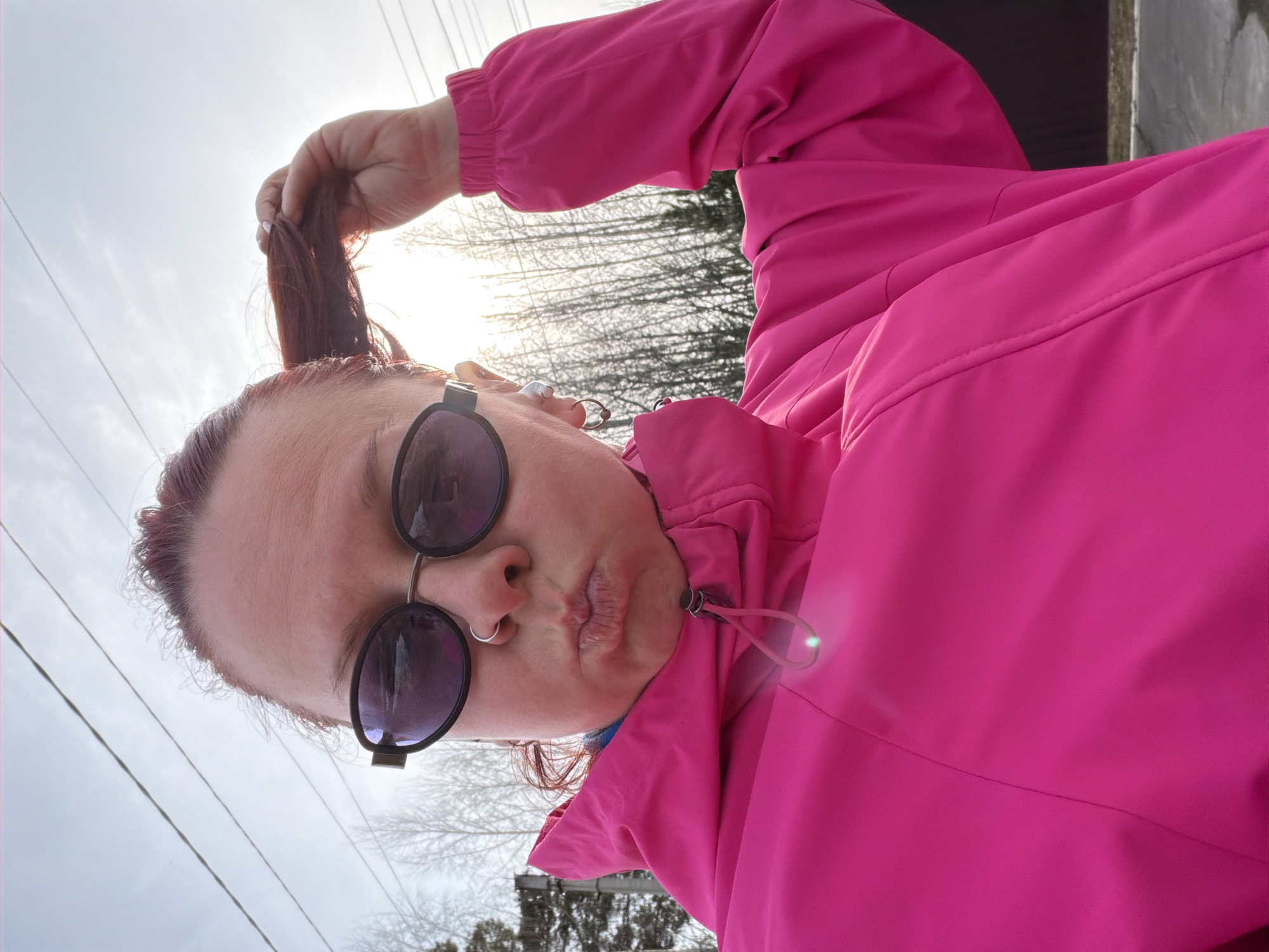 A woman is holding a portion of her hair and looking into the camera, puckering her lips, while standing outside on an overcast day in what appears to be Chaikovsky, Russia. She is wearing sunglasses and a bright pink jacket. In the background, there are bare trees and power lines.