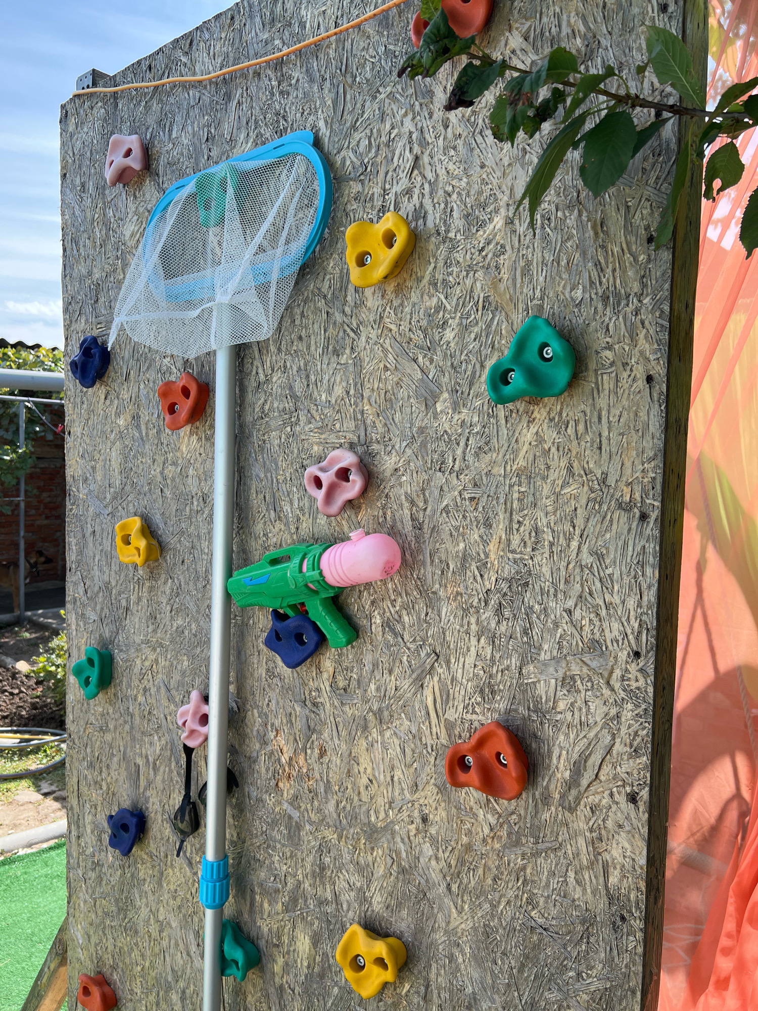 A climbing wall constructed from particleboard is outdoors, attached to a wooden frame. It is adorned with colorful plastic climbing holds in various shapes and sizes, predominantly red, yellow, green, blue, and pink. A blue-rimmed net on a long pole is attached to the wall, positioned near the top. Below the net, a green and pink toy water gun is mounted on the wall. A thin orange cable runs along the top of the wall. The background shows a blue sky with some clouds, foliage, and a hint of an orange tarp. The scene is located in the city of Morozovsk, Russia.