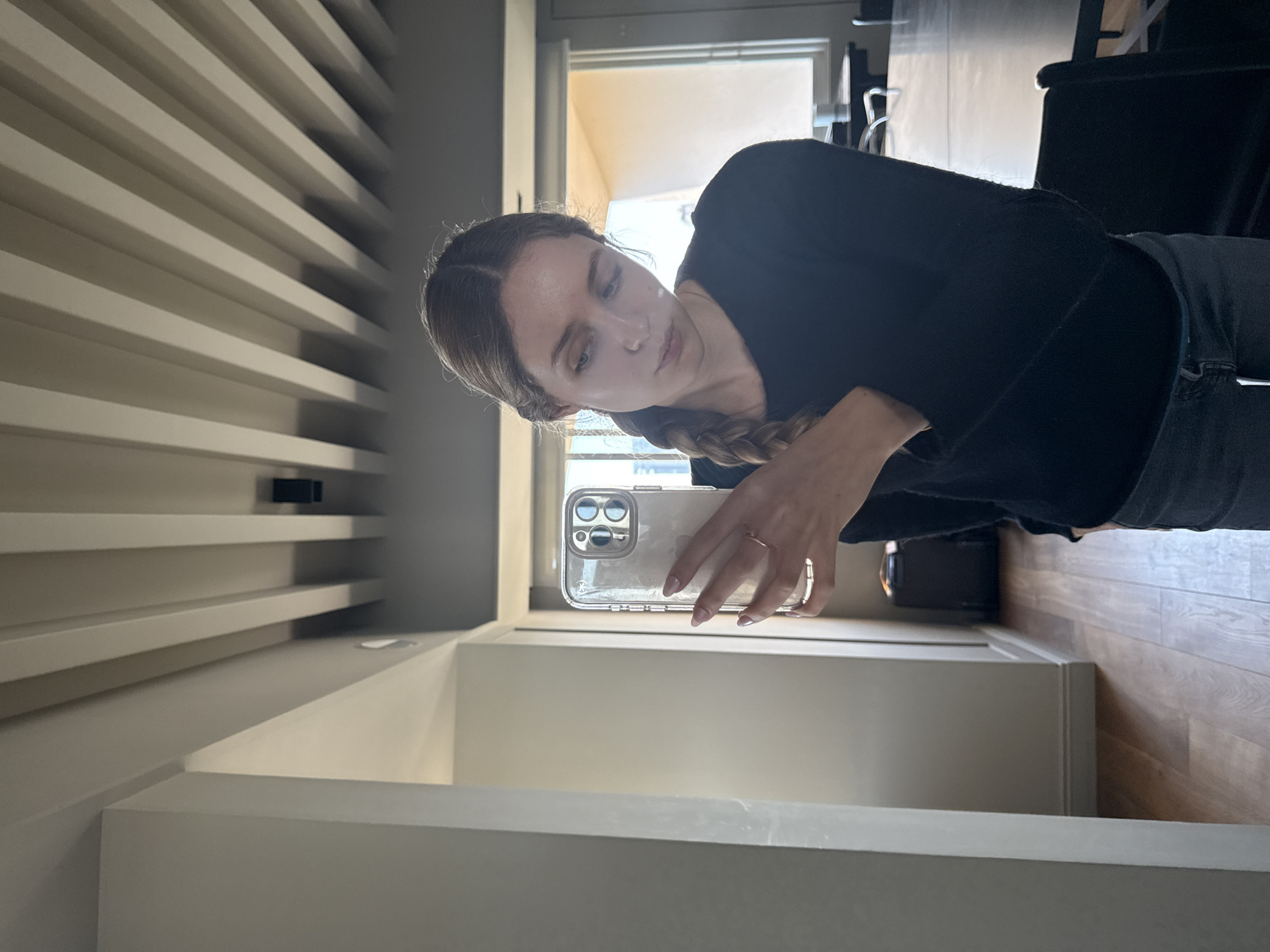 A young woman is depicted indoors, taking a mirror selfie or viewing her reflection in a smartphone. She has light brown hair styled in a braid over her left shoulder and wears a black long-sleeved top and dark jeans. Her gaze is directed downward towards the phone screen, giving her a contemplative expression. She holds a smartphone, likely an iPhone given the prominent triple camera module, encased in a clear protective case. A subtle "CASETiFY" logo is visible on the side of the phone case, and she wears a simple ring on the ring finger of her right hand, with light-colored nail polish on her fingernails.
The indoor setting features a modern aesthetic. To the left, a light-colored wall or room divider presents a series of horizontal slats or shelves, creating a textured background. A window is visible behind the woman, letting in bright natural light, indicating it is daytime. Elements of dark furniture or architectural features are faintly discernible through the window. The floor on the right side of the image appears to be made of light-colored wood. The overall ambiance is clean and contemporary.