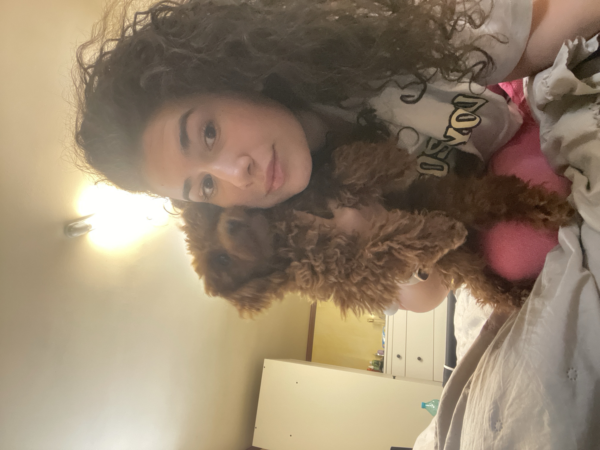 This image captures a young woman and a small, curly-haired brown dog in an intimate indoor setting, likely a bedroom in Sofia, Bulgaria. The woman, with dark curly hair, is lying down and looking towards the camera with a gentle, slightly smiling expression. She is cuddling or holding the small dog, which has a rich reddish-brown coat and appears calm and affectionate, resting its head close to her. A bright pink, spherical object, possibly a dog toy, is visible near the dog's body.
The woman is wearing a light-colored t-shirt with partial text visible on it, appearing to say "DO YO". The background features light-colored walls and bedding, suggesting a cozy environment. A bright artificial light source, possibly a ceiling light, casts a warm glow from the upper left portion of the frame (when correctly oriented). To the right, a white wardrobe or cabinet with dark handles can be seen, indicating typical home furnishings. The overall atmosphere conveys comfort, warmth, and affection between the woman and her pet. The indoor lighting suggests it could be evening or night.