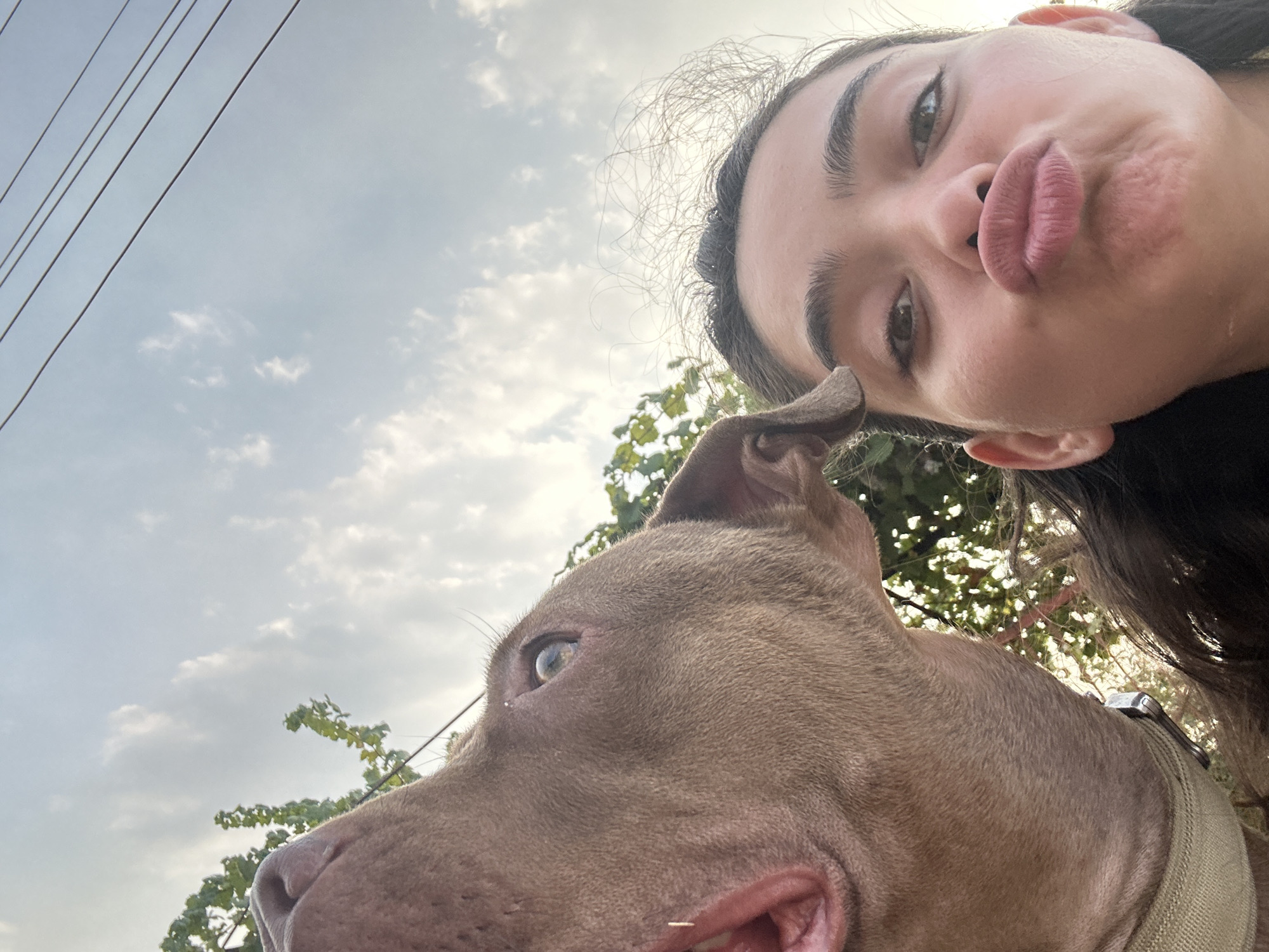 The image, taken outdoors with a cloudy sky and power lines overhead, features a selfie of a young woman and a dog. The woman, positioned on the right side of the frame, is looking directly at the camera with her lips puckered in a kissing gesture. Her eyebrows are dark and well-defined, and she has dark hair. To her left, a large dog, likely a pit bull type breed, is prominently featured, its head angled towards the camera and its mouth slightly open, revealing its tongue and teeth. The dog appears to be nudging its head towards the woman. Green foliage from trees is visible in the background, suggesting a natural or semi-urban setting. The photo's low angle and close-up perspective create an intimate and playful atmosphere. The location is identified as Malevo, Bulgaria.