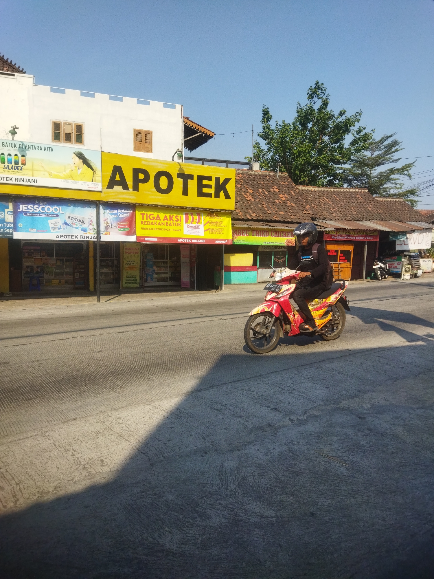 A medium shot captures a street scene in Juwiring, Indonesia, under a clear, bright blue sky. A man wearing a black helmet and dark clothing rides a red and yellow motorcycle with elaborate graphics, moving from right to left across the frame. He is slightly blurred, indicating motion.
The motorcycle is on a paved road with distinct parallel lines etched into its surface, suggesting it's a well-traveled area. The road is partly in shadow, cast by an unseen object to the left, creating a strong contrast.
To the left of the road, a row of buildings with white and yellow facades lines the street. A prominent yellow sign with large black lettering reads "APOTEK" (Pharmacy). Below this, various shop fronts are visible, displaying products and signs in Indonesian. One sign reads "JESSCOOL" with images of cooling products. Another advertises "TIGA AKSI REDAKAN BATUK" (Three Actions Relieve Cough).
The buildings have tiled roofs, typical of Indonesian architecture. Behind the shops, a lush green tree rises, its branches reaching towards the sky. A power line is visible against the blue. Further to the right, more buildings with corrugated metal roofs and wooden elements can be seen, with a glimpse of a sign that appears to be for a food establishment, possibly a fried chicken place given the text "FRIED CHICKEN" visible on one section. A parked motorcycle is also visible in front of one of these buildings.
The overall impression is of a commercial street in a town or city in Indonesia, bustling with everyday activity.