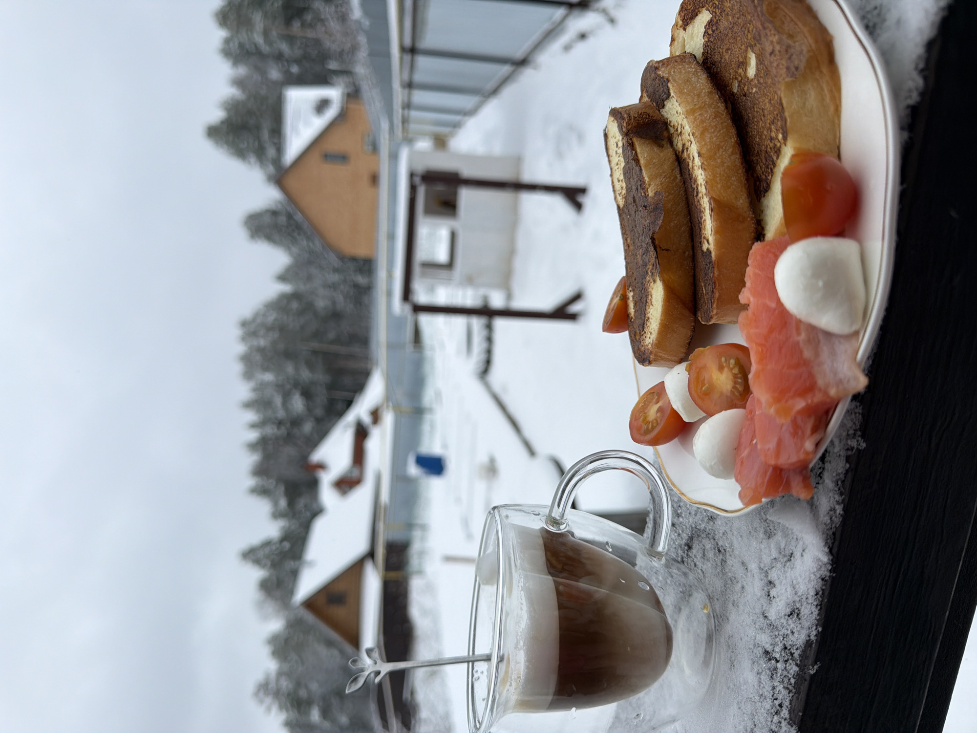 A breakfast scene is captured on what appears to be a balcony or outdoor space covered in snow. In the foreground, a glass mug filled with a coffee drink rests next to a white plate. The plate holds sliced bread, cherry tomatoes, mozzarella balls, and salmon. The background shows a winter landscape with houses and trees, suggesting a residential area in a snowy region. The image is taken at an angle, enhancing the perspective of the outdoor setting. Given the location specified as Chaikovsky, Russia, the scene is indicative of a winter morning in a Russian town.