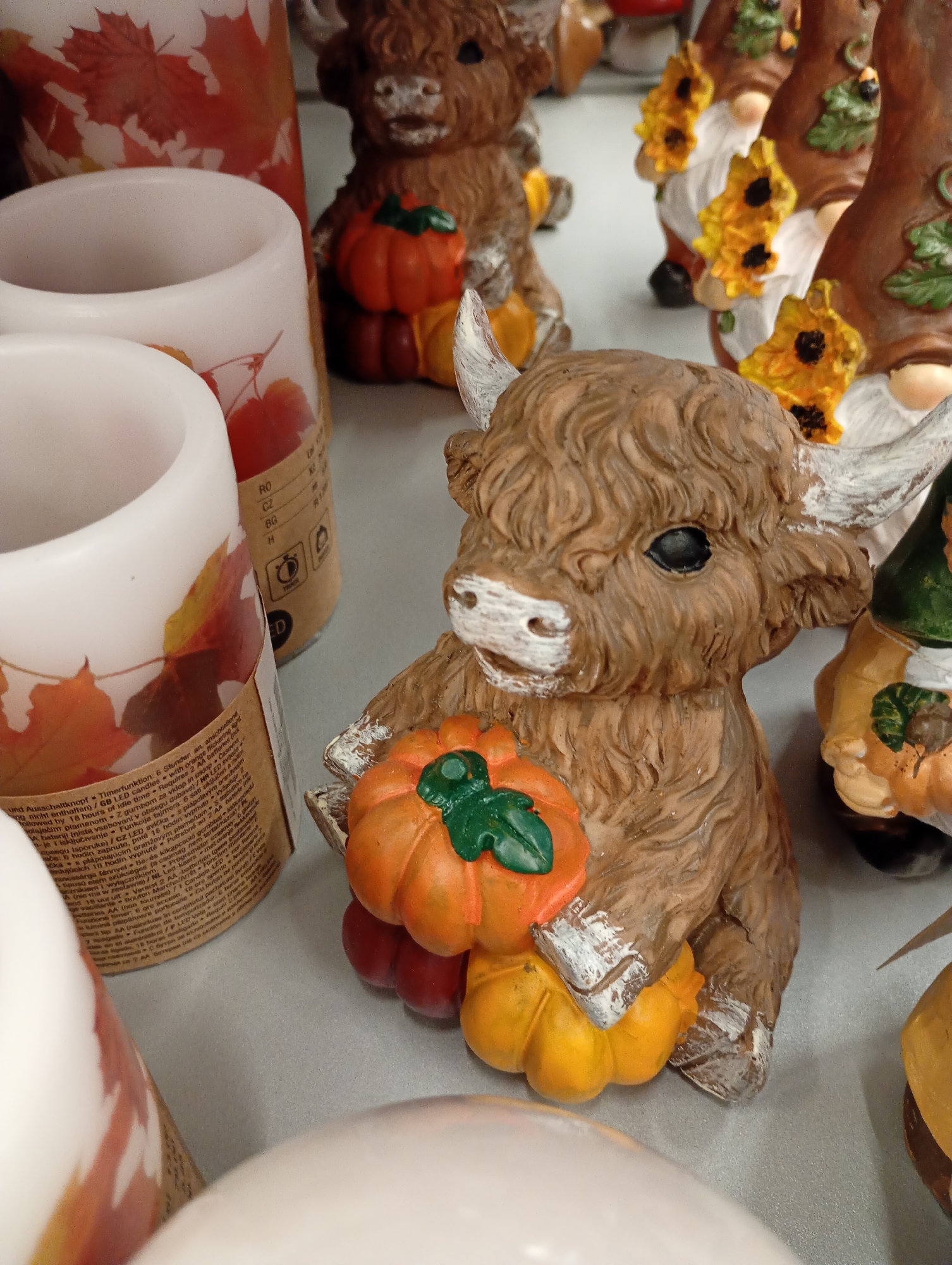 The image displays a close-up of decorative items, likely for sale at a retail establishment. In the foreground, a brown, fuzzy highland cow figurine is prominently featured, holding two small pumpkins, one orange and one yellow. The cow has white accents on its nose and hooves. Behind the cow, to the left, are two white, cylindrical candles decorated with autumn leaves. To the right of the cow, several gnome figurines are visible, adorned with sunflowers and leaves. The background is out of focus, suggesting a shelf or display in a store. The overall scene evokes an autumnal or harvest theme. The items are positioned on a flat, gray surface, likely a shelf in a store. There is no indication of the specific location within the city of Bad Lauterberg im Harz, Germany, beyond the general context of a retail environment.