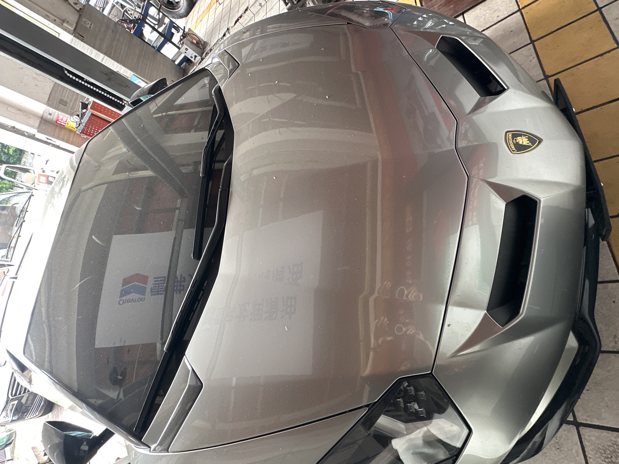 A silver Lamborghini Aventador is parked in what appears to be a service area or garage, possibly at a dealership or repair shop. The car's hood is prominently featured, showing a reflection on its surface. A portion of a logo with the text "LAMBORGHINI" is visible on the front bumper. In the background, there are hints of other vehicles, industrial equipment, and structural elements that suggest an urban, commercial, or industrial setting. The floor is tiled in a checkered pattern of yellow and grey. The lighting appears to be artificial, consistent with an indoor environment. The scene is captured from a slightly elevated, forward-facing perspective.