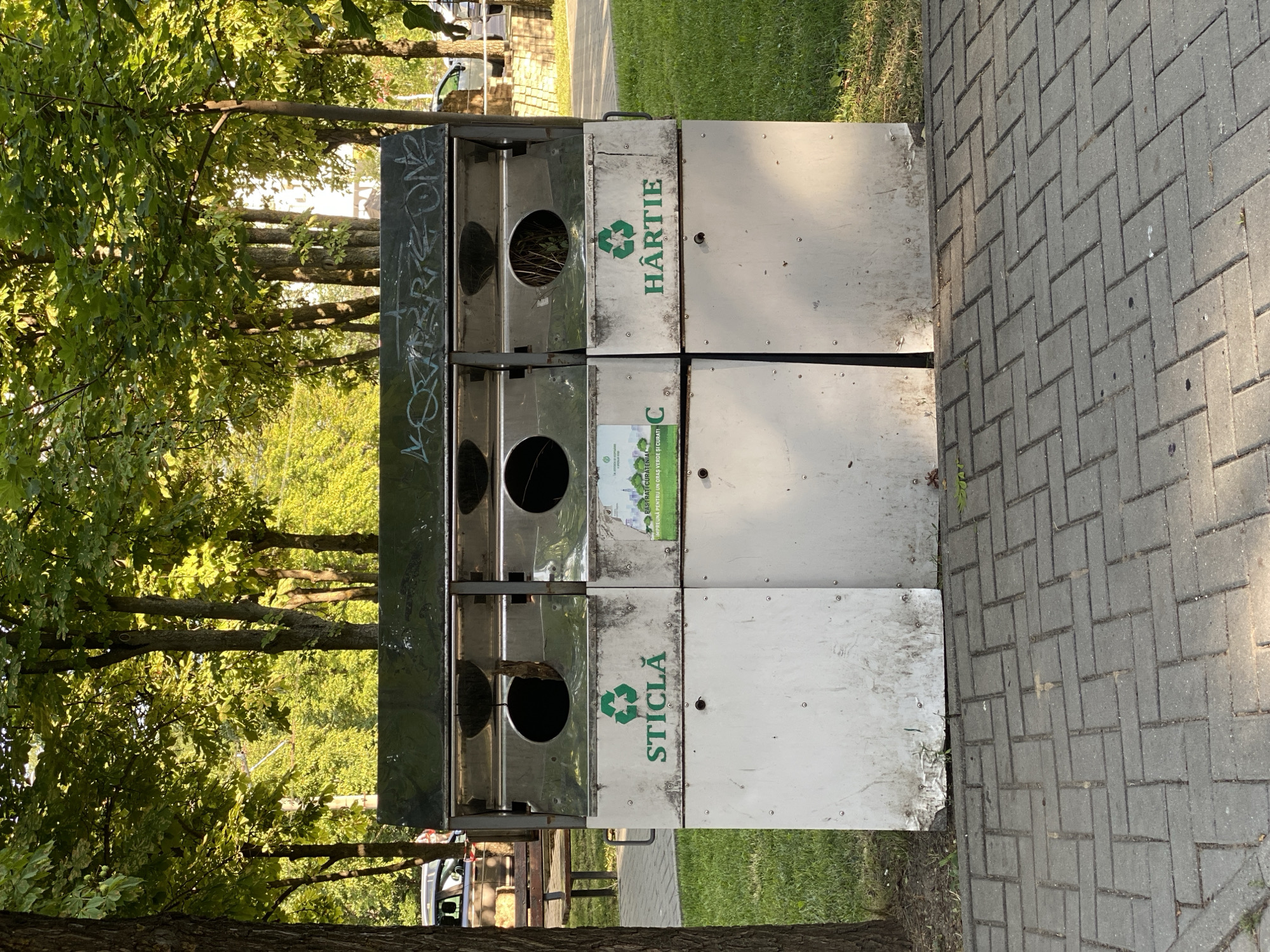 The image depicts a recycling station with three compartments in a public park in Chișinău, Moldova. The recycling station is made of metal, with green recycling symbols and labels indicating the types of waste to be deposited: "STICLĂ" (glass) on the left, and "HÂRTIE" (paper) on the right. The middle compartment has a smaller label with an image of trees and text that reads "STRĂLUCIȚI CURĂȚENIA IMPREUNĂ UN ORAȘ VERDE ȘI CURAT!" (Shine the cleanliness together, a green and clean city!) and "C" in a circle indicating it's for other recyclables, possibly plastic or general recycling. The recycling station is situated on a paved walkway made of grey interlocking bricks, with a grassy area and trees in the background. The sun is shining, casting shadows from the trees. A wooden bench is visible to the left of the recycling station, and a car is parked in the distance. The overall scene suggests a well-maintained urban park.