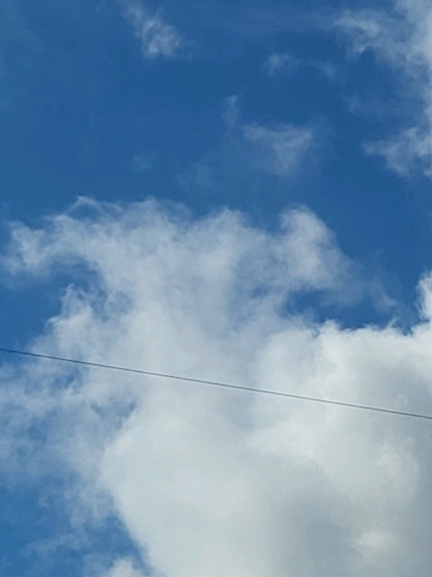 A bright blue sky with scattered white clouds is visible. A thin, dark horizontal line, likely a power line or wire, stretches across the lower middle portion of the frame. The image composition suggests an outdoor scene, possibly taken from the ground looking upwards. There are no people or specific landmarks that would definitively place the image in Jelgava, Latvia, or any other specific urban location. The focus is solely on the sky and a single wire.