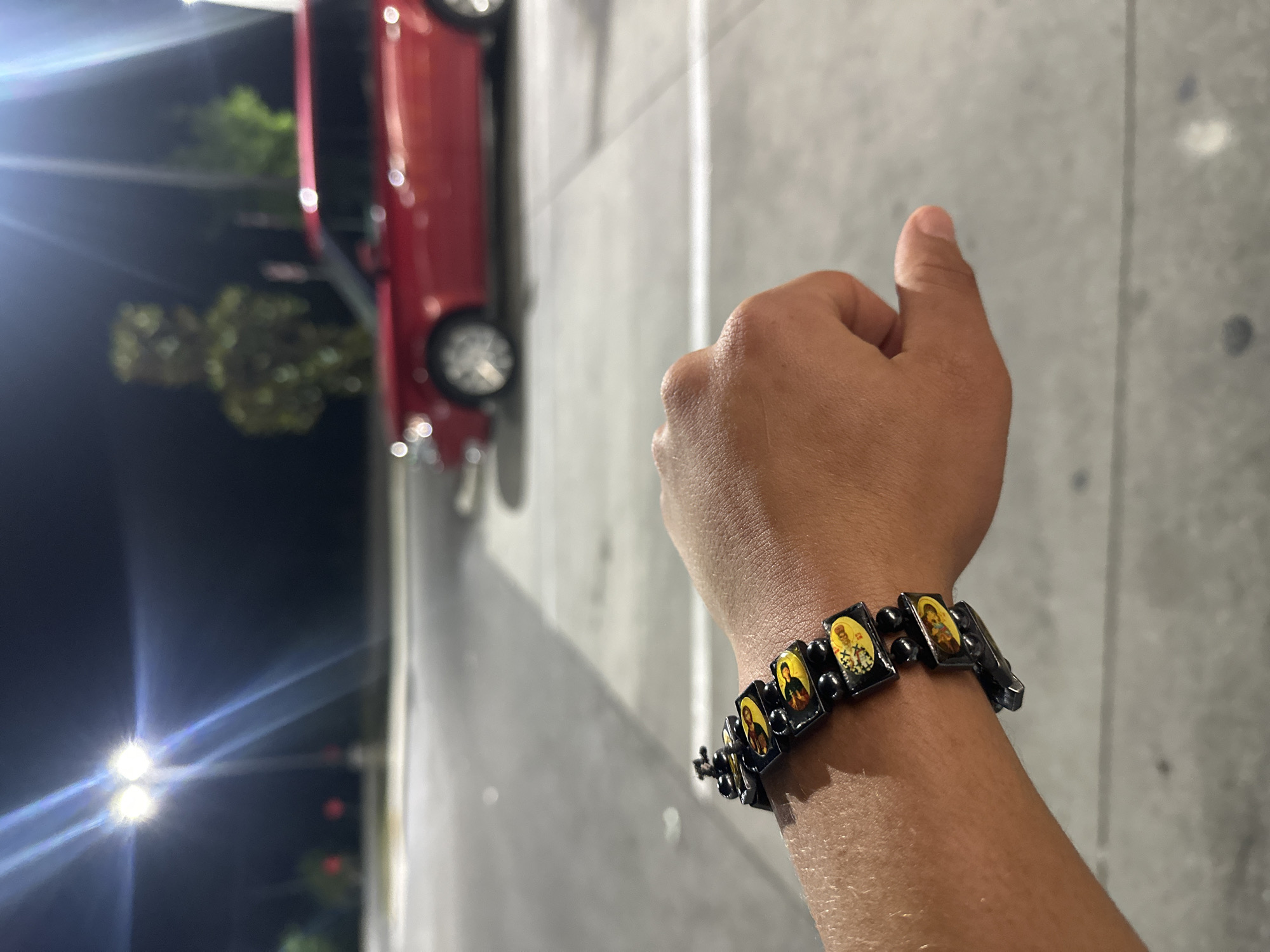 A close-up, outdoor shot shows a person's right hand and wrist with a black bracelet featuring small, rectangular images of religious figures against a yellow background. The person's thumb is extended upwards. In the blurred background, a red vehicle is visible, with a dark sky, streetlights, and greenery further in the background. The location cannot be determined as Homosassa Springs.