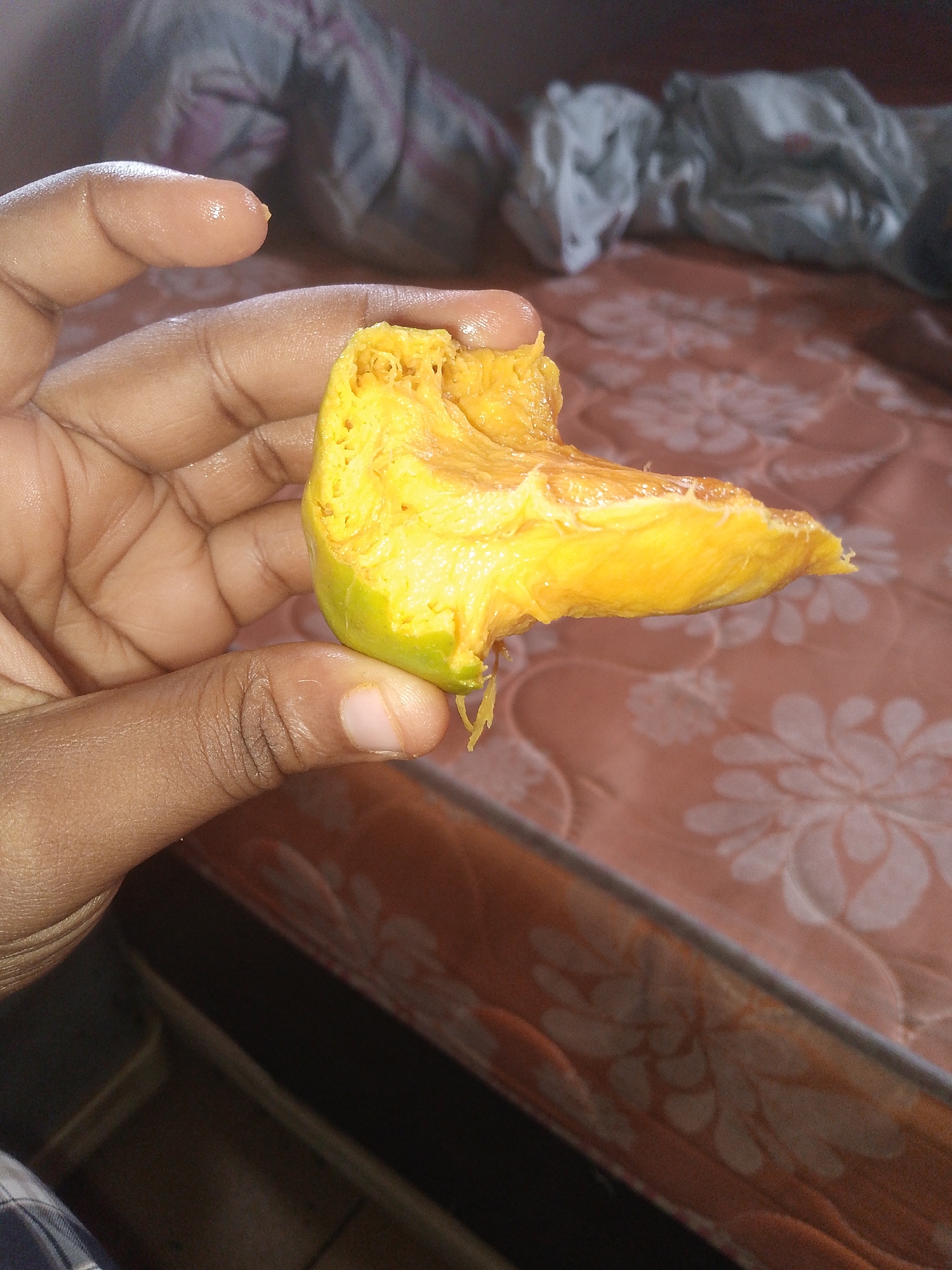 A dark-skinned hand holds a section of a yellow-orange fruit with green skin, possibly a mango, indoors. The background includes a bed with a floral pattern and a crumpled white cloth. The location is Unknown, Unknown.