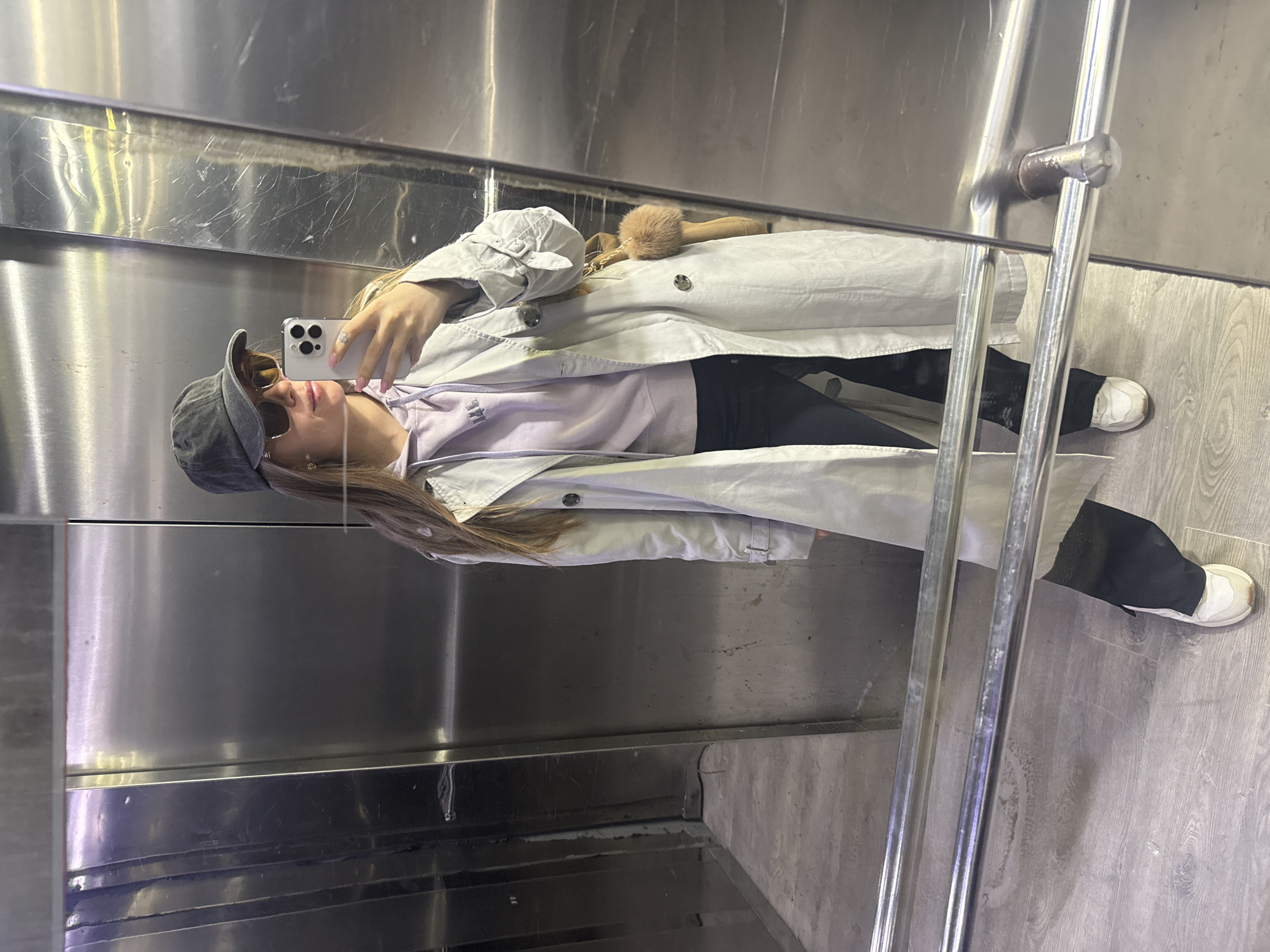 A woman is taking a selfie in what appears to be an elevator. She is wearing a gray baseball cap, sunglasses, a lavender sweater, black pants, a light-colored trench coat, and white sneakers. She is holding a smartphone in her right hand, facing the mirror of the elevator. The elevator has stainless steel walls and a metal railing. The floor has a wood-grain pattern. The location is possibly in the city of Batumi, Georgia.