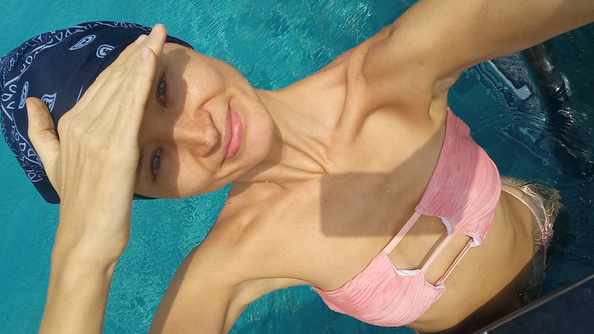 A woman wearing a dark blue bandana on her head and a pink bikini is in a swimming pool. Her right hand is held up near her forehead. The water is turquoise in color. The scene appears to be in daylight. The location is in Pattaya City, Thailand.
