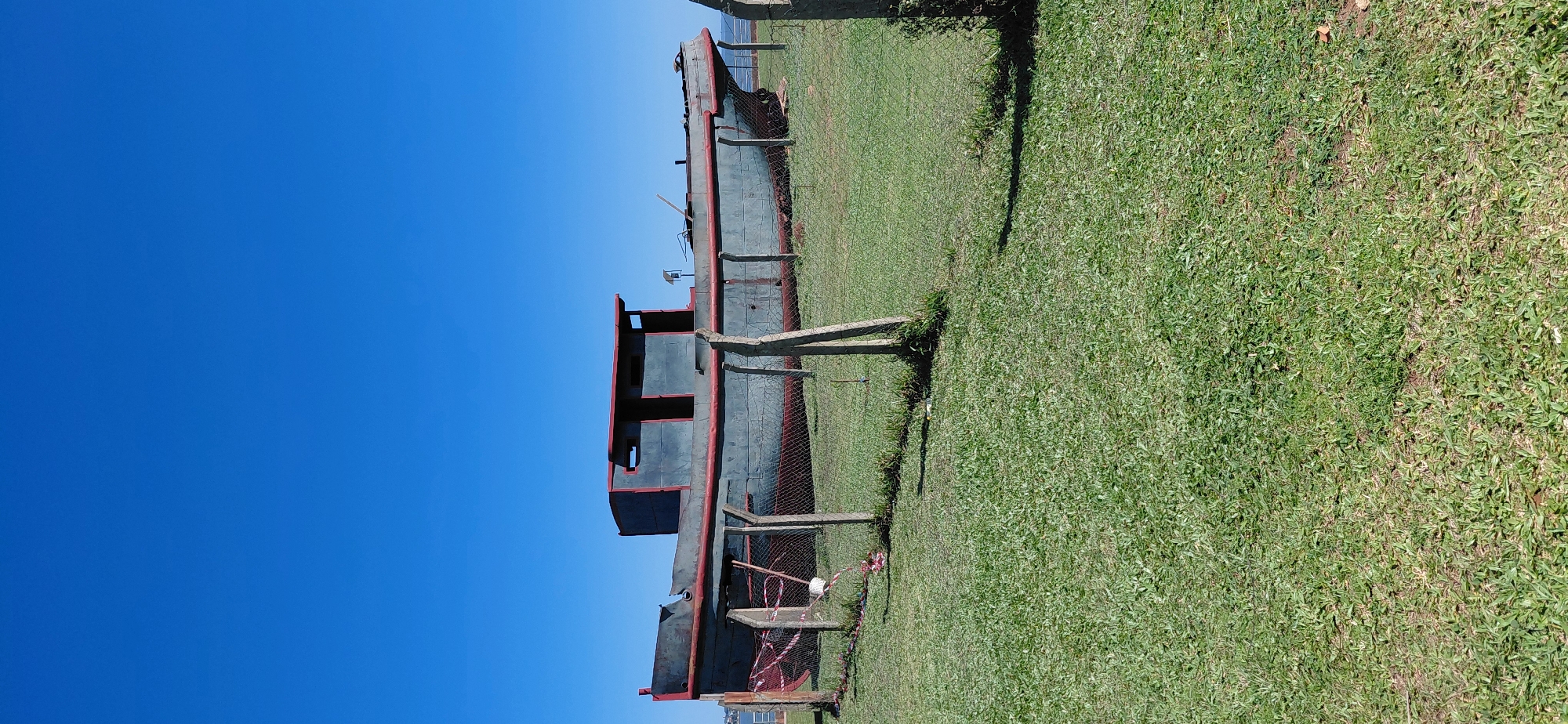 An old, disused boat, painted gray with a red trim, rests on a grassy area behind a chain-link fence. The boat has a weathered appearance and a cabin structure on its deck. The fence is supported by concrete posts and has some red and white caution tape attached to it. The surrounding area is a well-maintained lawn, and in the distance, a body of water and a clear blue sky are visible. This scene is located in the city of Encarnacion, Paraguay, likely a park or public space near the water. There are no people visible in the image.
