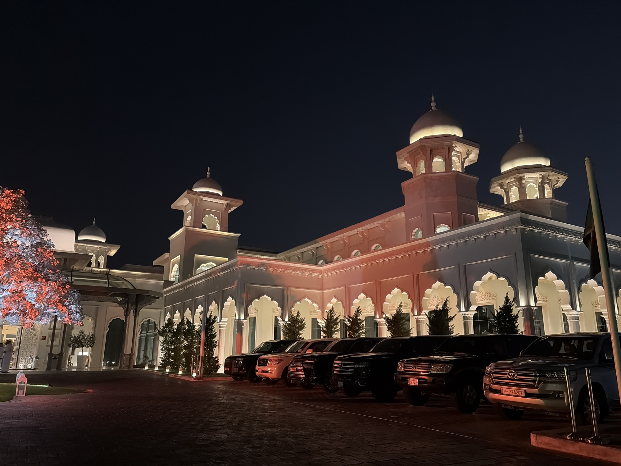 This is a nighttime, eye-level, medium shot of a white building with distinctive architectural features, possibly a hotel or government building, in an outdoor setting. The building is illuminated with warm-toned lights, highlighting its arched windows and multiple domed towers. In front of the building, a row of several black and silver luxury SUVs are parked facing the viewer. The foreground is a paved area with a cobblestone-like pattern. To the left of the building, a tree is decorated with red and white lights. The sky above is dark, indicating it is night. No people are clearly visible in the scene. The license plates on the vehicles are in Arabic. The scene is serene and orderly, with a sense of grandeur.