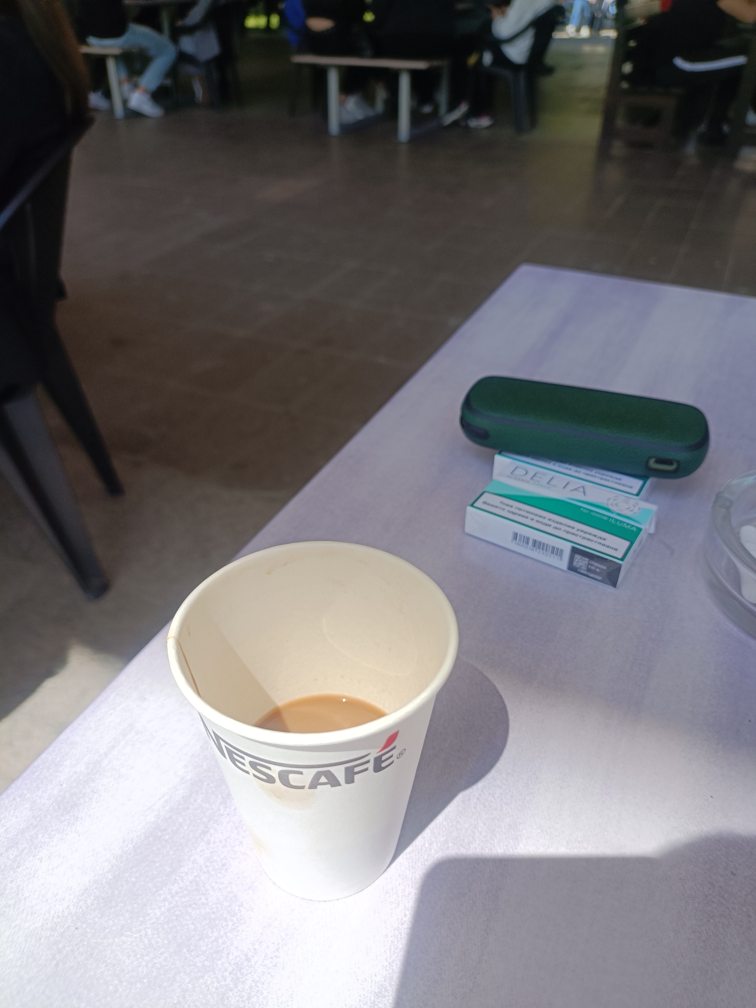 The image presents a close-up view of a table set in an outdoor or semi-outdoor public area, likely a café or communal seating space. Strong shadows cast by objects on the table indicate bright daylight and sunny weather.
In the foreground, a white paper cup, prominently featuring the "NESCAFÉ" logo, holds a partially consumed, light brown beverage, likely coffee with milk. To the right of the cup, on the light-colored table surface, several items are arranged. These include a dark green, oblong case, possibly for an electronic device, and two small white and teal boxes. The boxes are labeled "DELIA" and specify "for use in IQOS ILUMA," identifying them as tobacco sticks. Visible Cyrillic text on the packaging translates to "smoke-free tobacco product" and "Keep away from children," further detailing their nature. A small portion of a clear ashtray is also visible on the far right.
The background is softly blurred, showing indistinct figures of people seated at other tables and dark chair legs, suggesting a casual gathering or a moment of relaxation. The overall scene conveys a laid-back atmosphere. The specific tobacco product packaging is consistent with the provided location context of Plovdiv, Bulgaria.