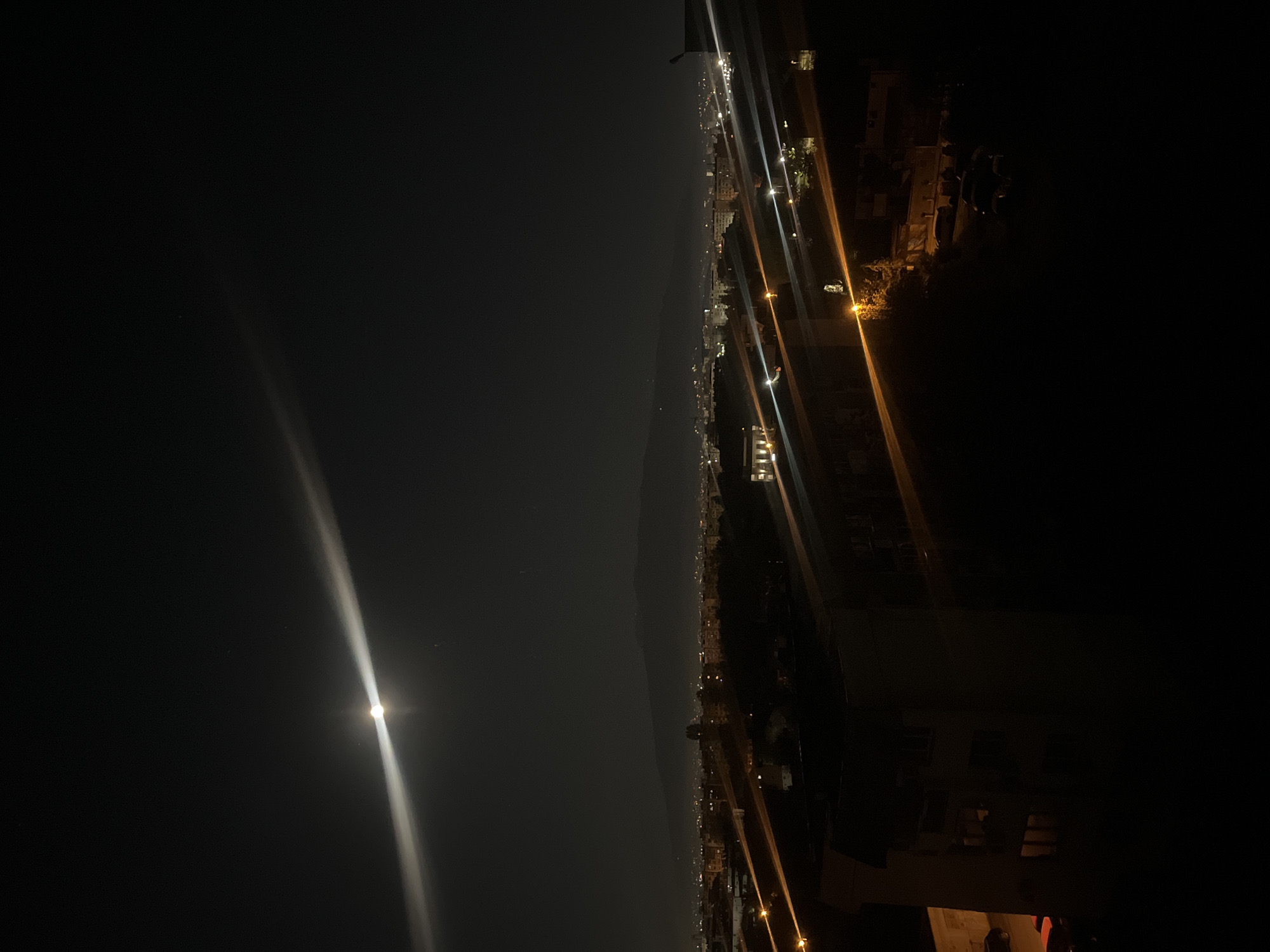 The image shows a nighttime view of Sofia, Bulgaria, with the full moon visible in the left portion of the frame. A strip of buildings and lights of the city run across the middle of the image. There are streetlights and some buildings, although they are mostly in darkness. The overall lighting is dim. The photo appears to be taken from a high vantage point, possibly a balcony or rooftop.