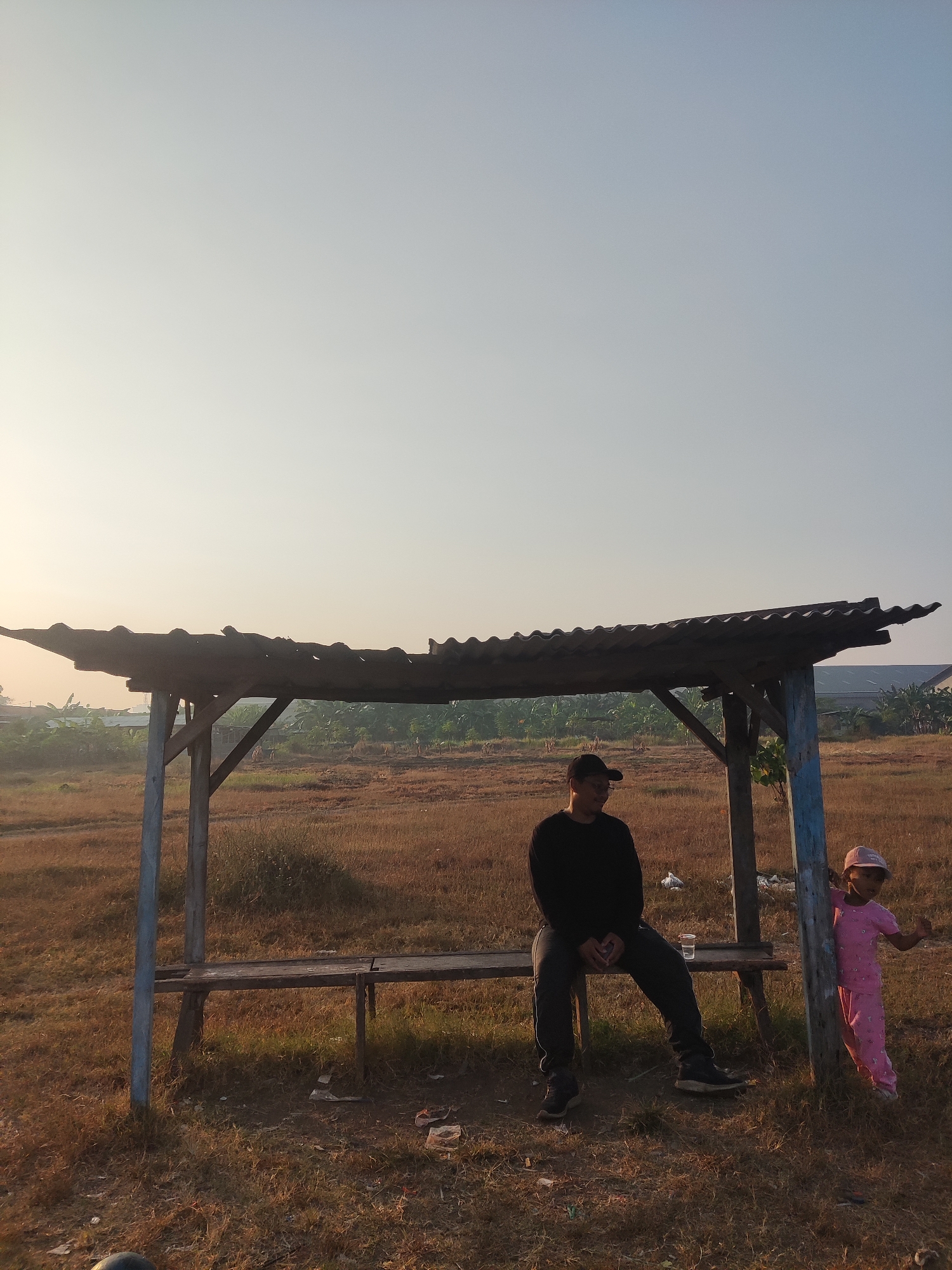 In Banjarkemantren, Indonesia, under a simple, dilapidated open-air shelter with a corrugated roof, a man and a young child are present. The man, wearing dark clothing and a baseball cap, is seated on a wooden bench. To his right, a child dressed in pink pajamas and a pink cap stands near a support post of the shelter. The shelter is situated in a dry, grassy field with sparse vegetation. In the background, a line of trees and some structures are visible under a hazy, pale sky, suggesting it is either early morning or late afternoon. The overall scene appears to be a rural or semi-rural outdoor setting.