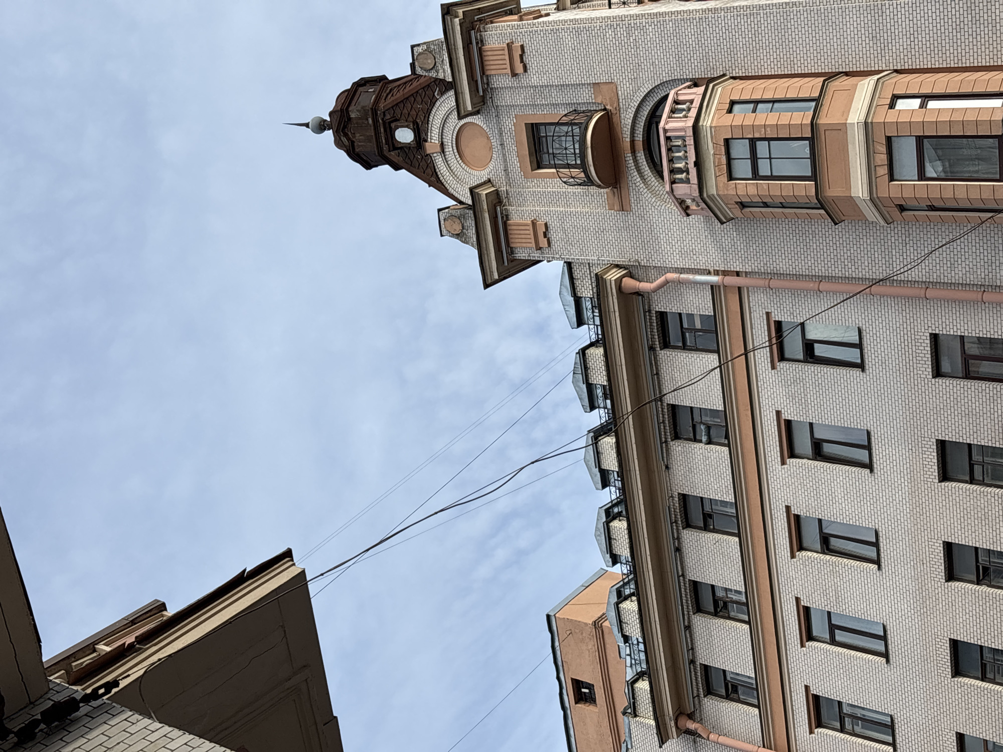 A low-angle, upward-looking photograph, significantly rotated clockwise, captures the facades of two historic buildings against an overcast sky in what appears to be Saint Petersburg, Russia.
The primary building, occupying the right and upper portions of the rotated frame, is a tall, ornate structure constructed with light-colored brickwork. It features prominent reddish-brown architectural accents, including decorative bands, cornices, and window surrounds. The building is topped by an elaborate turret or dome, constructed from weathered, possibly copper or zinc, metal, culminating in a pointed spire with a spherical finial. Below the turret, there are intricate details such as a circular decorative medallion and an arched opening. The facade also includes a decorative rounded balcony with a black metal railing and a multi-faceted bay window section. Numerous rectangular windows are visible across its surface, and a light-colored drainpipe runs vertically.
A second building is partially visible in the lower-left corner, showcasing a simpler, light-colored facade and a flatter roofline. The sky is a uniform, pale blue with a few faint, wispy clouds, indicating a daytime scene, likely under overcast conditions. Several thin, dark utility wires crisscross the expanse of the sky between the two buildings. The architectural style of the main building, with its decorative brickwork, distinctive turret, and detailed bay windows, is characteristic of historic European architecture often found in cities like Saint Petersburg. There is no visible text in the image.