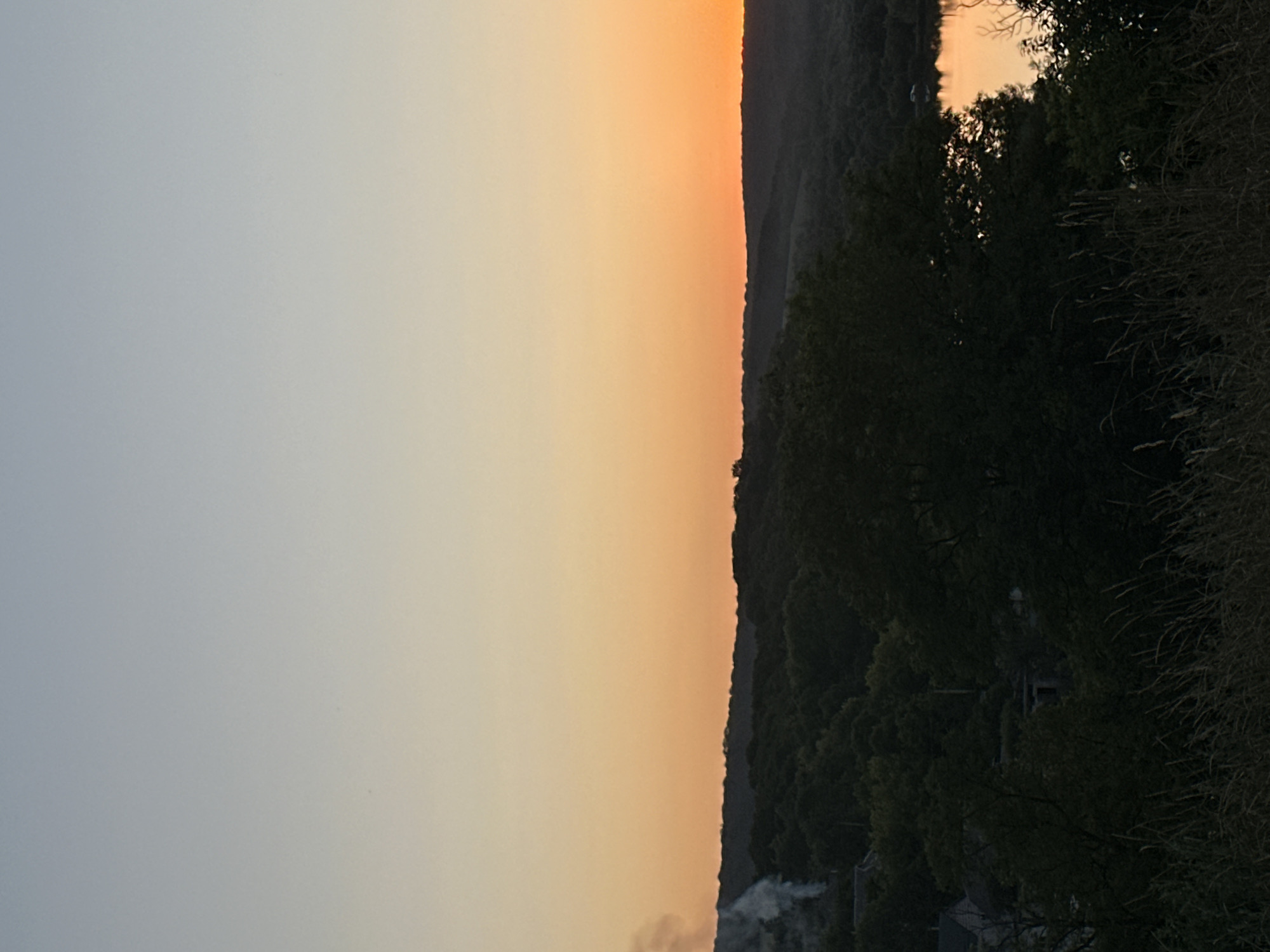 The image depicts a panoramic view of a natural landscape at dusk, potentially near Makiv, Ukraine. The sky is a gradient of pale blue to warm orange, indicating the sun is low on the horizon. A dense forest covers the rolling hills in the mid-ground, with a body of water, likely a lake or river, reflecting the orange hues of the sunset on the right side of the frame. In the far left, a plume of white smoke rises from behind the treeline, suggesting human activity, possibly a controlled burn or a chimney. Some structures, possibly houses, are faintly visible nestled amongst the trees on the left. The foreground is filled with tall, dry grasses, silhouetted against the darker landscape. No people are visible in the image.