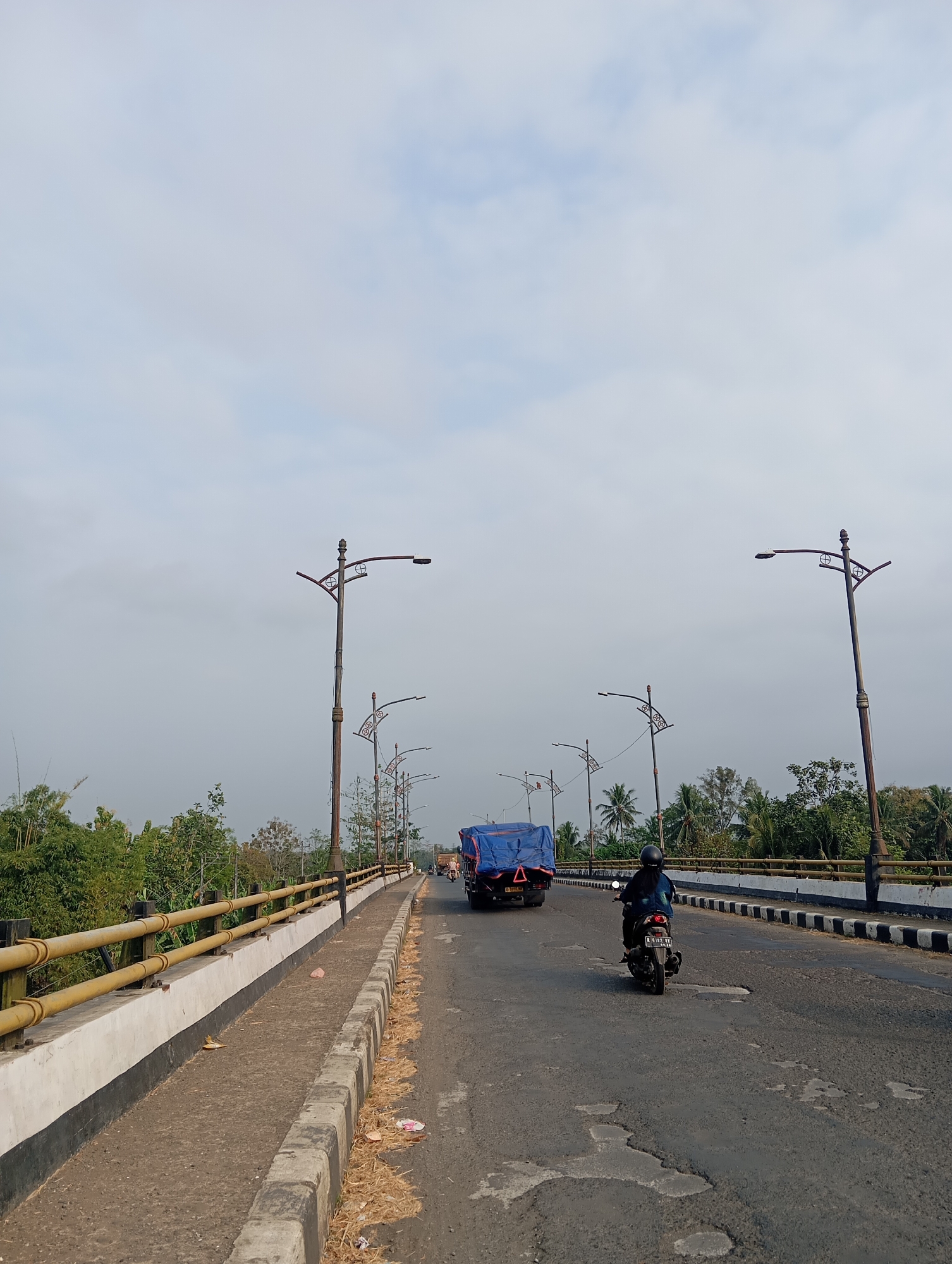 The image captures a street scene on a bridge in Kedungbenda, Indonesia. The road, which appears to be in fair condition with some visible potholes, is flanked by a pedestrian walkway on the left and a guardrail. Several ornate lampposts line the bridge, casting their shadows on the asphalt. In the foreground, a person rides a motorcycle, and further down the road, a truck covered with a blue tarp is visible. Lush green trees and vegetation can be seen on either side of the bridge, with a hazy sky overhead.