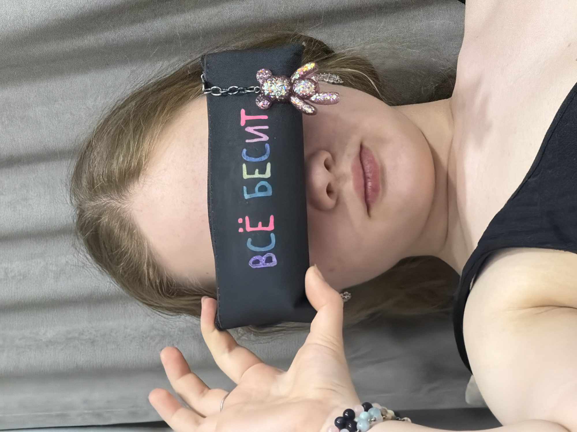A pale-skinned person with light brown hair is lying on a grey surface. A black blindfold with a chain and a sparkly bear adornment covers their eyes. The blindfold has the word "ВСЁ БЕСИТ" written vertically on it in multi-colored letters. The person is wearing a black top. Their hand, adorned with a bracelet of black and light blue beads, is holding the blindfold. The background is a plain grey. There is no information available in the image to determine the location as Chaikovsky, Russia.