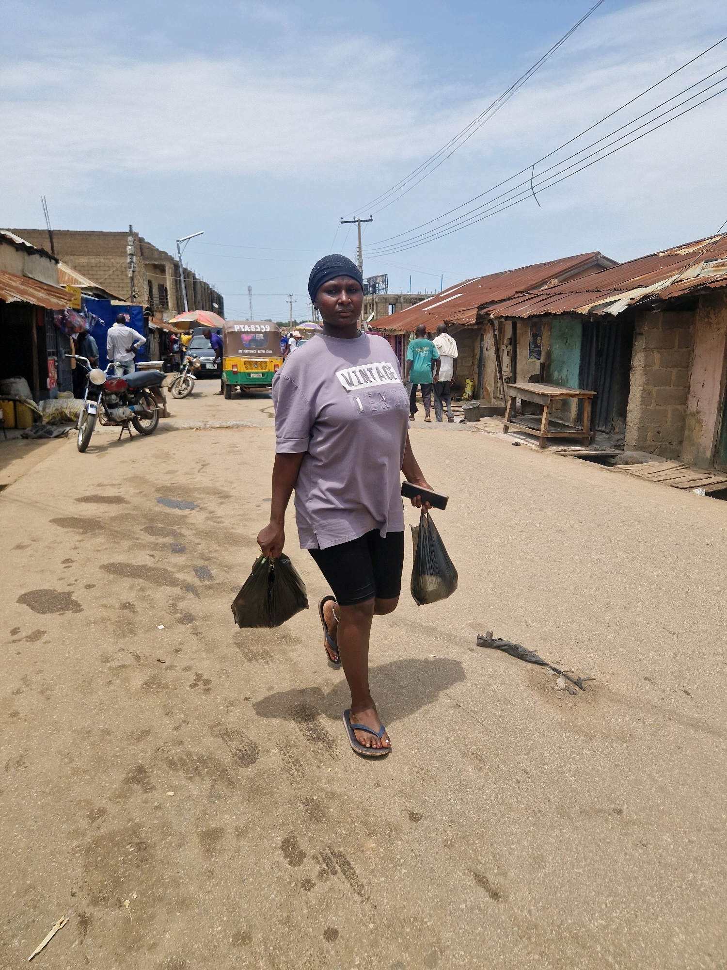 A woman walks down a dusty street in an outdoor market. She is wearing a purple t-shirt with "VINTAGE" printed on it and black shorts. She carries two black plastic bags, one in each hand, and a black phone in her right hand. Her feet are bare, except for blue flip-flops. In the background, several stalls line the street, with corrugated tin roofs. A yellow auto-rickshaw with "PTA-8339" on it is parked in the middle ground, along with a motorcycle and other vehicles. The sky is partly cloudy. The ground is a mix of dirt and some scattered debris.