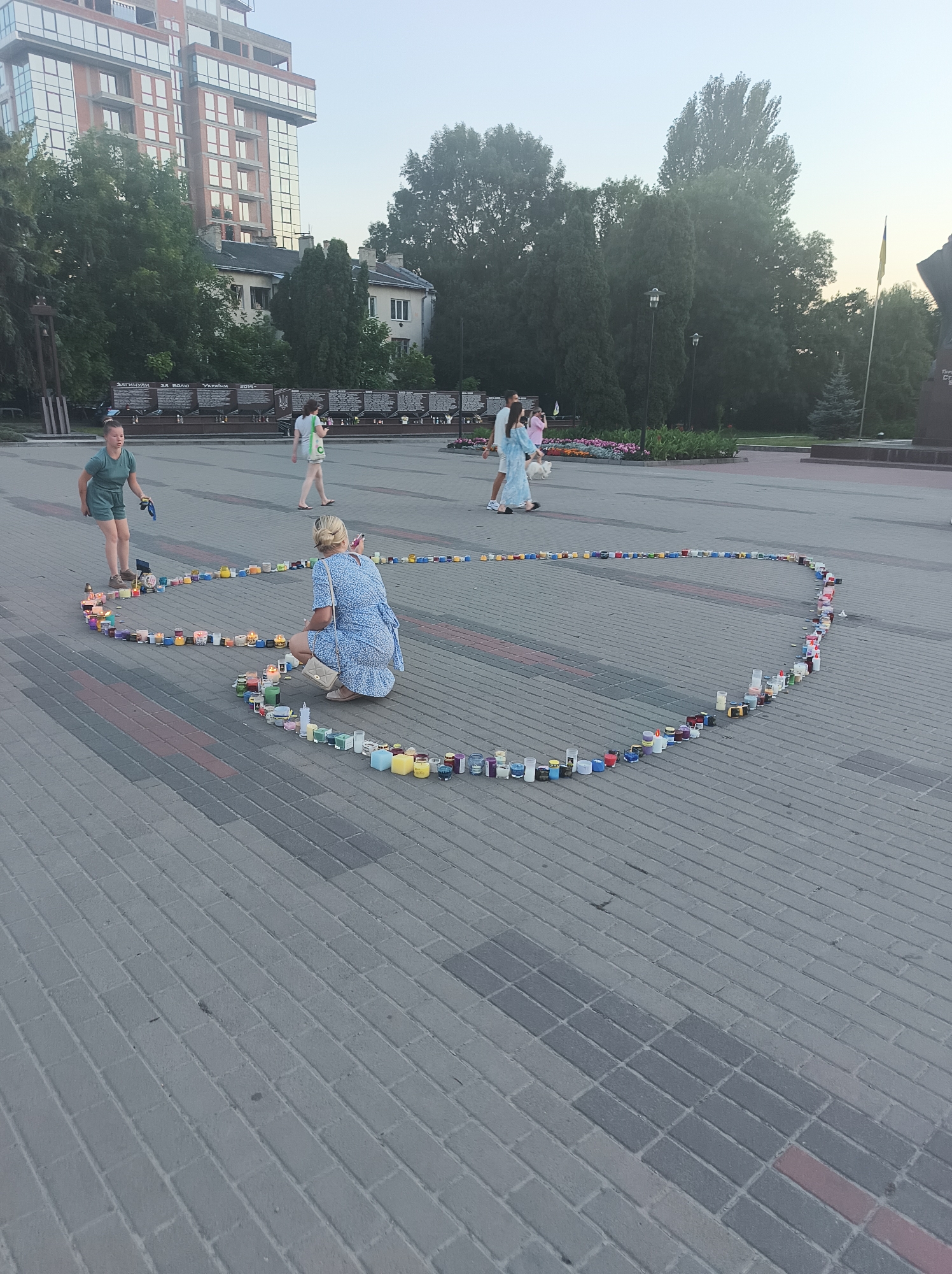 The image depicts an outdoor scene in Ternopil, Ukraine, likely a public square or park, with a focus on a heart-shaped arrangement of lit candles on a paved surface. In the foreground, a woman in a blue dress is kneeling and observing the candles, holding a smartphone. Another woman, wearing a green t-shirt and shorts, stands to the left, also looking at the candles. Several other people are visible in the background, including a couple walking with a small dog.
The environment includes a modern building with many windows in the upper left, mature trees, and a Ukrainian flag visible in the distance. A monument or memorial with inscriptions is also present in the background. The ground is paved with grey interlocking bricks. The lighting suggests it is either early morning or late evening. The arrangement of candles appears to be a memorial or a form of public display.